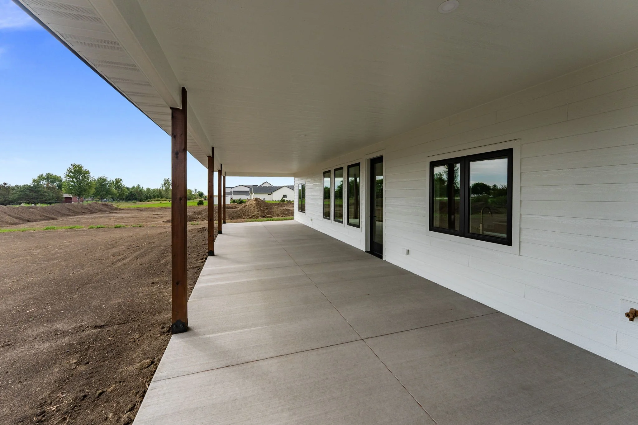 Newly built house with a white exterior, black-framed windows, a covered patio with concrete flooring, and wooden support posts, situated in a suburban area with open land and neighboring houses in the background.