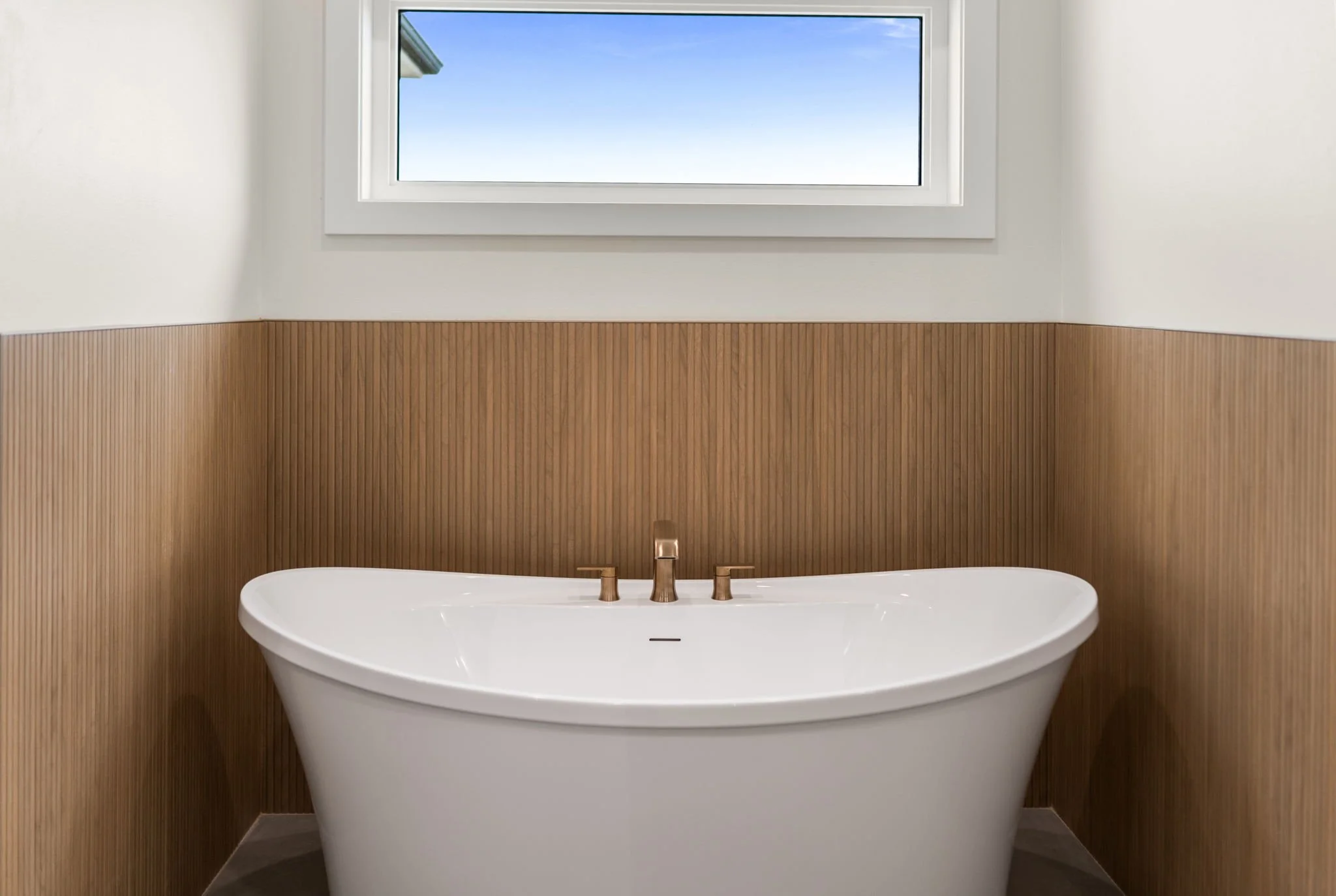 A modern bathroom with a freestanding bathtub, a wooden accent wall behind it, a small rectangular window letting in natural light, and a golden faucet.