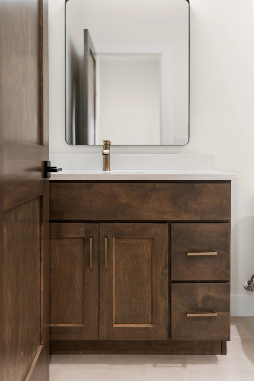 A bathroom vanity with a wooden cabinet, white countertop, and a brass faucet, reflected in a wall-mounted mirror.