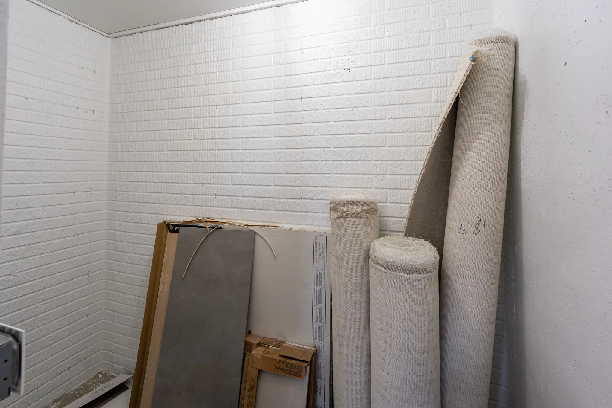 Corner of a room with white brick textured walls partially covered with various rolls of wallpaper or mural paper, some leaning against the wall, and some in boxes.
