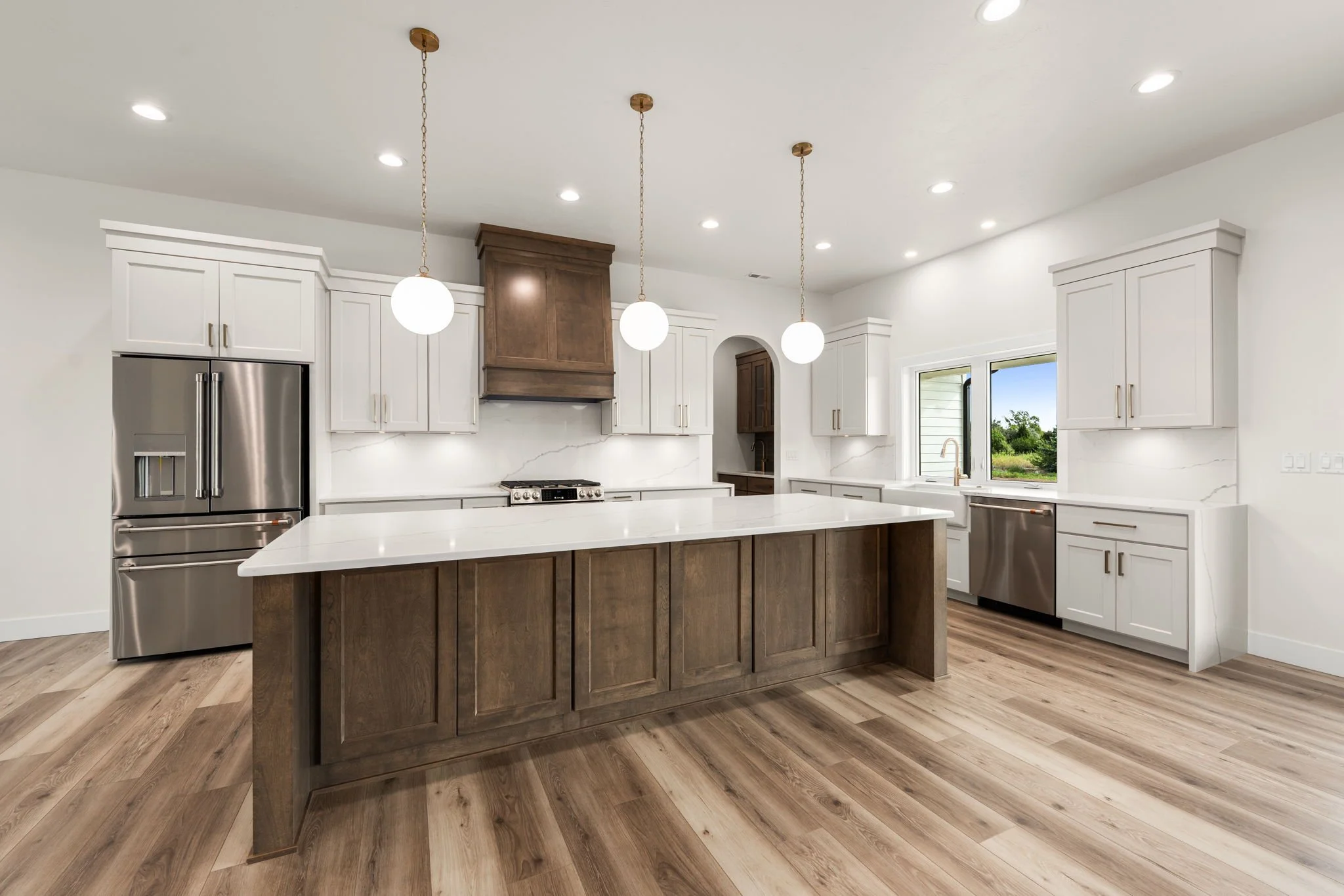 Modern kitchen with white cabinets, a large kitchen island with a wooden base, stainless steel appliances, and a window revealing outdoors. Three pendant lights hang above the island, and the kitchen has wood flooring.