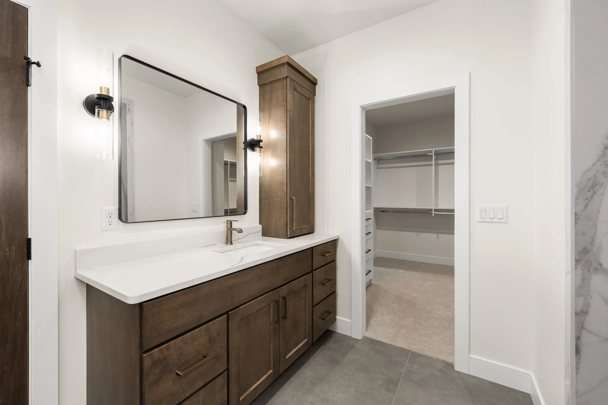 Modern bathroom vanity with a large rectangular mirror, a dark wood cabinet, and a white marble countertop with a brass faucet. An adjoining walk-in closet with white shelving is visible in the background.
