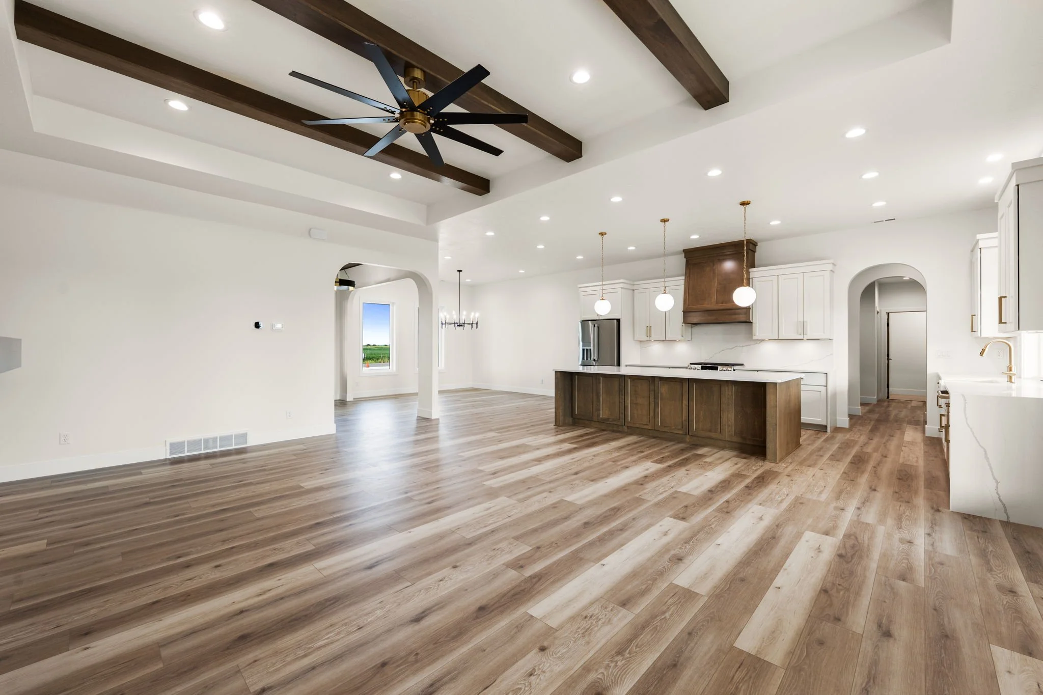 Open-concept kitchen and living area with wood flooring, white walls, a dark wood ceiling beam, recessed lighting, a ceiling fan, pendant lights, white cabinets, and a stainless steel refrigerator.