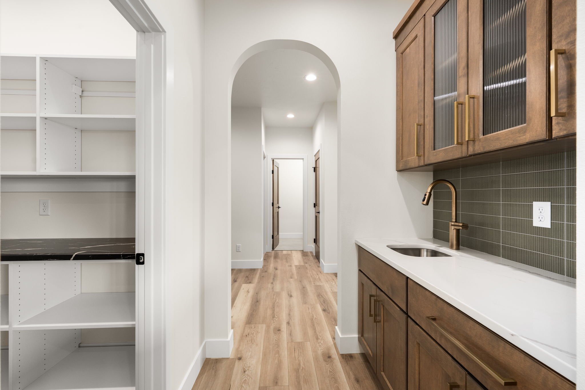 Interior of a modern kitchen with wooden cabinets, a white countertop, a gold faucet, and gray backsplash tiles. There is an open pantry with white shelves on the left and a hallway with multiple doors ahead.