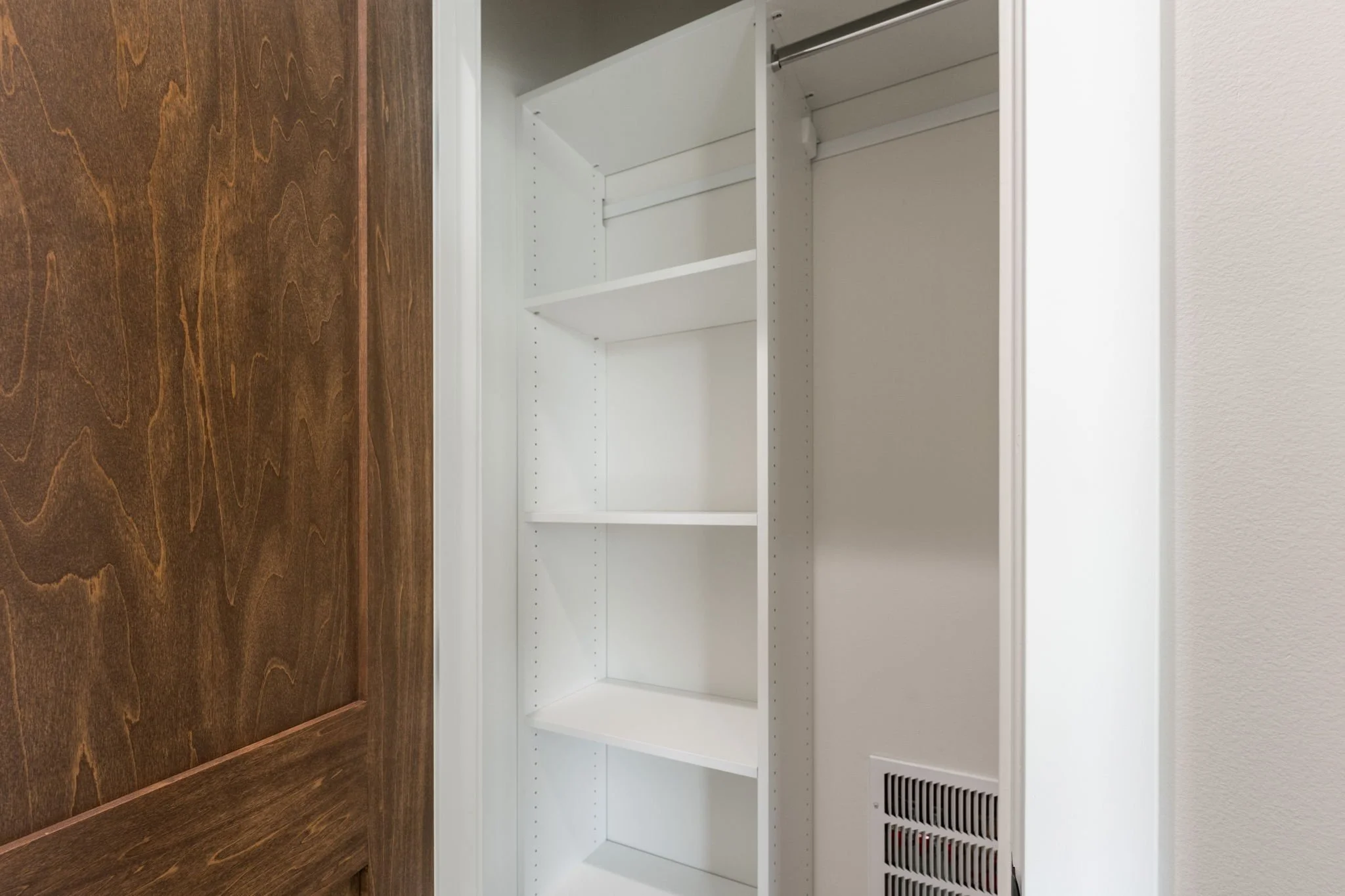 Empty white built-in kitchen cabinet or pantry with shelves and a vent at the bottom, adjacent to a wood-paneled wall and a white textured wall.