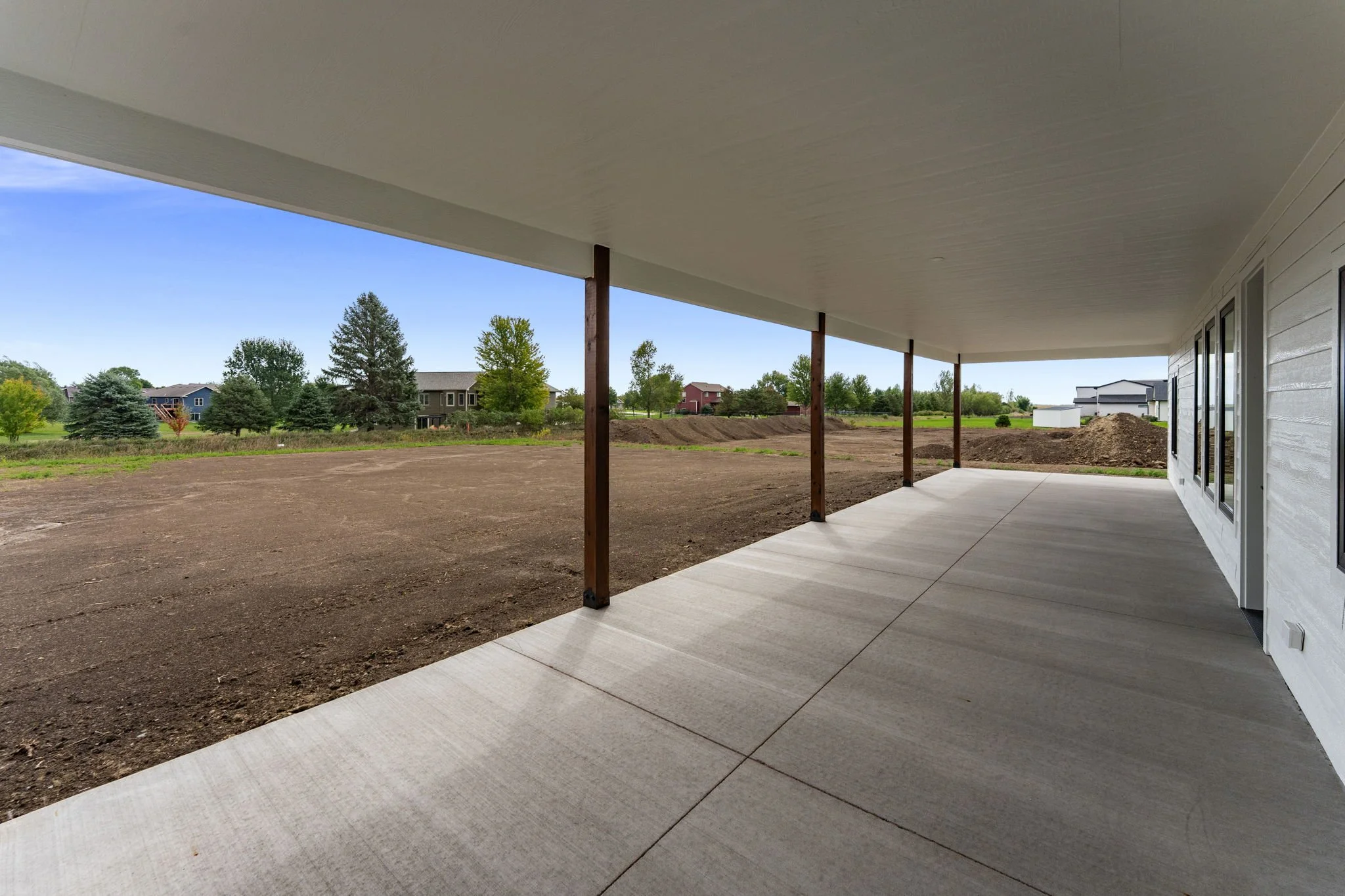 View of a spacious outdoor patio with a concrete floor and a white ceiling supported by wooden posts, overlooking a landscaped yard with trees and neighboring houses under a clear blue sky.