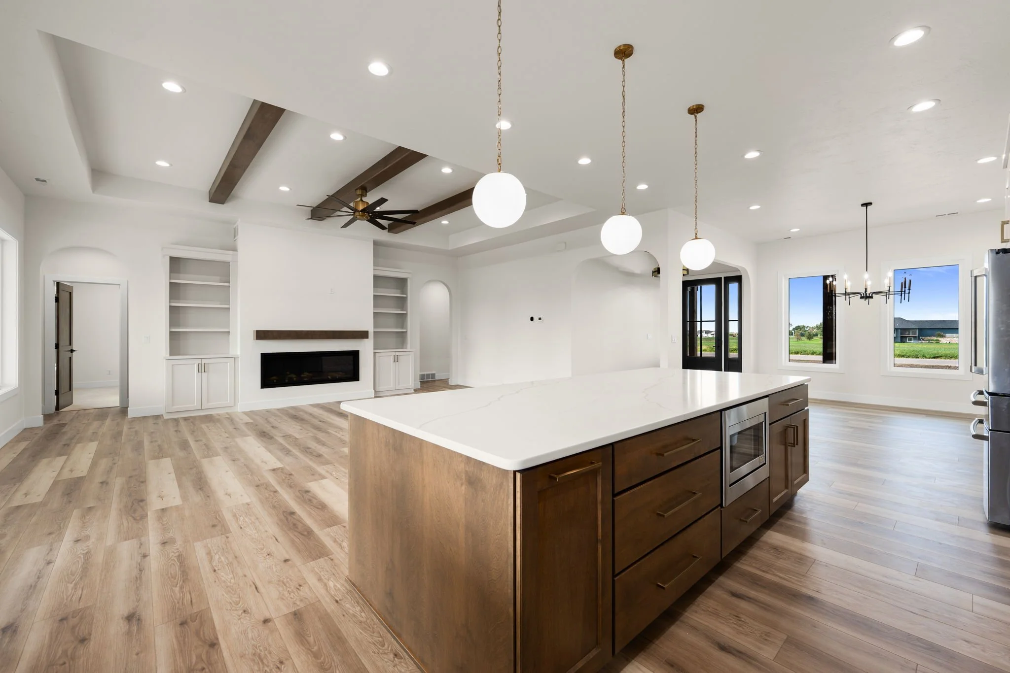 Modern kitchen with a large kitchen island, pendant lights, white walls, and hardwood floors. There are large windows and double black doors leading outside.