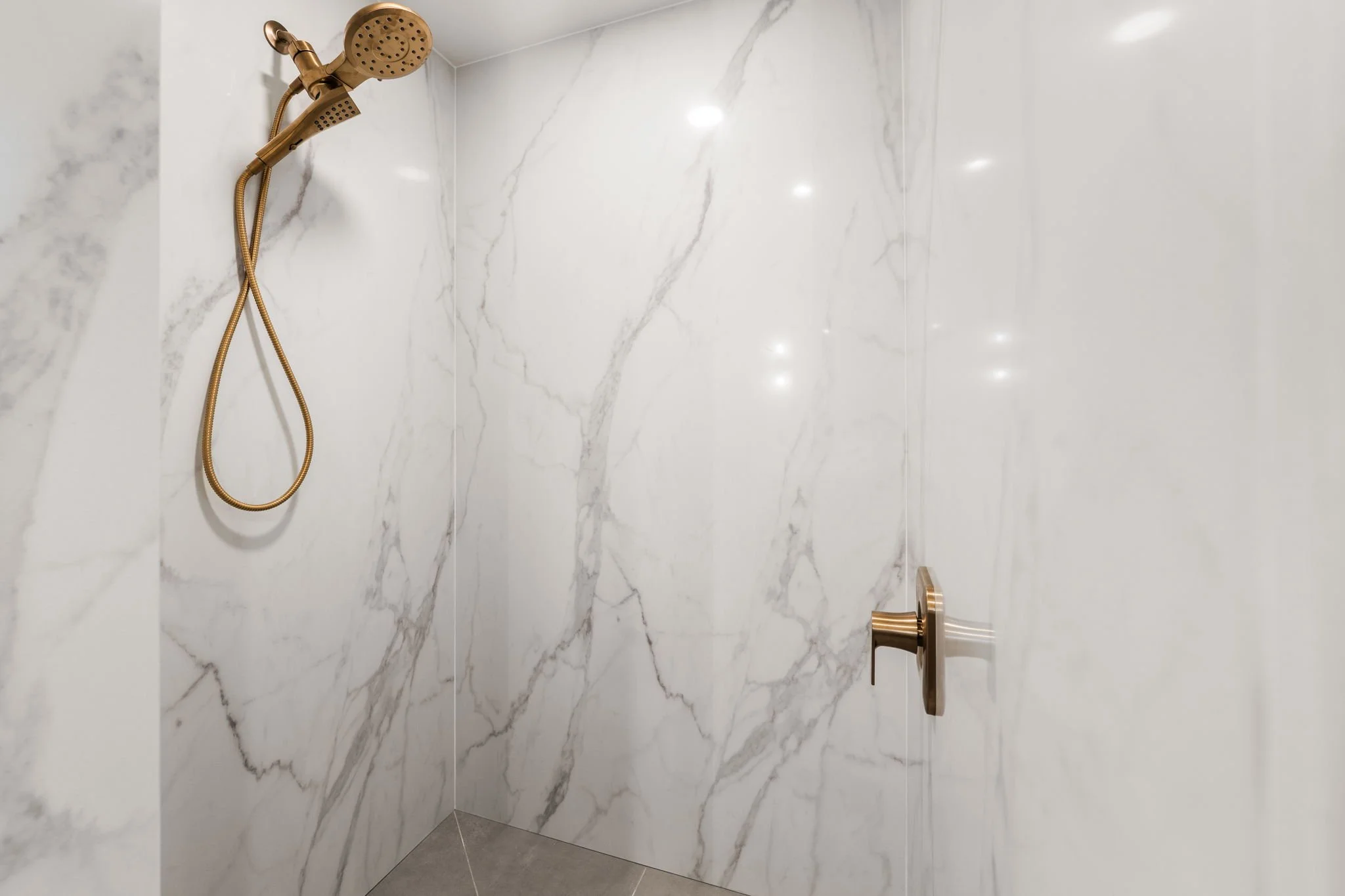 Shower area with white marble walls featuring gray veining, a gold showerhead, and a matching gold handle.