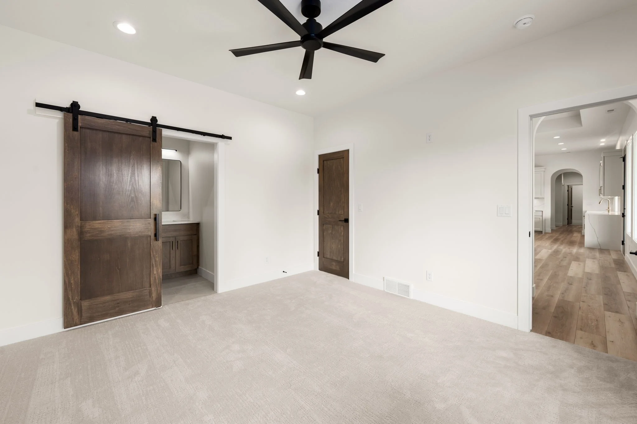 Interior of a modern house with white walls, a beige carpeted floor, a ceiling fan, and a sliding barn door made of dark wood. A doorway reveals a hallway with hardwood floors and white cabinetry.