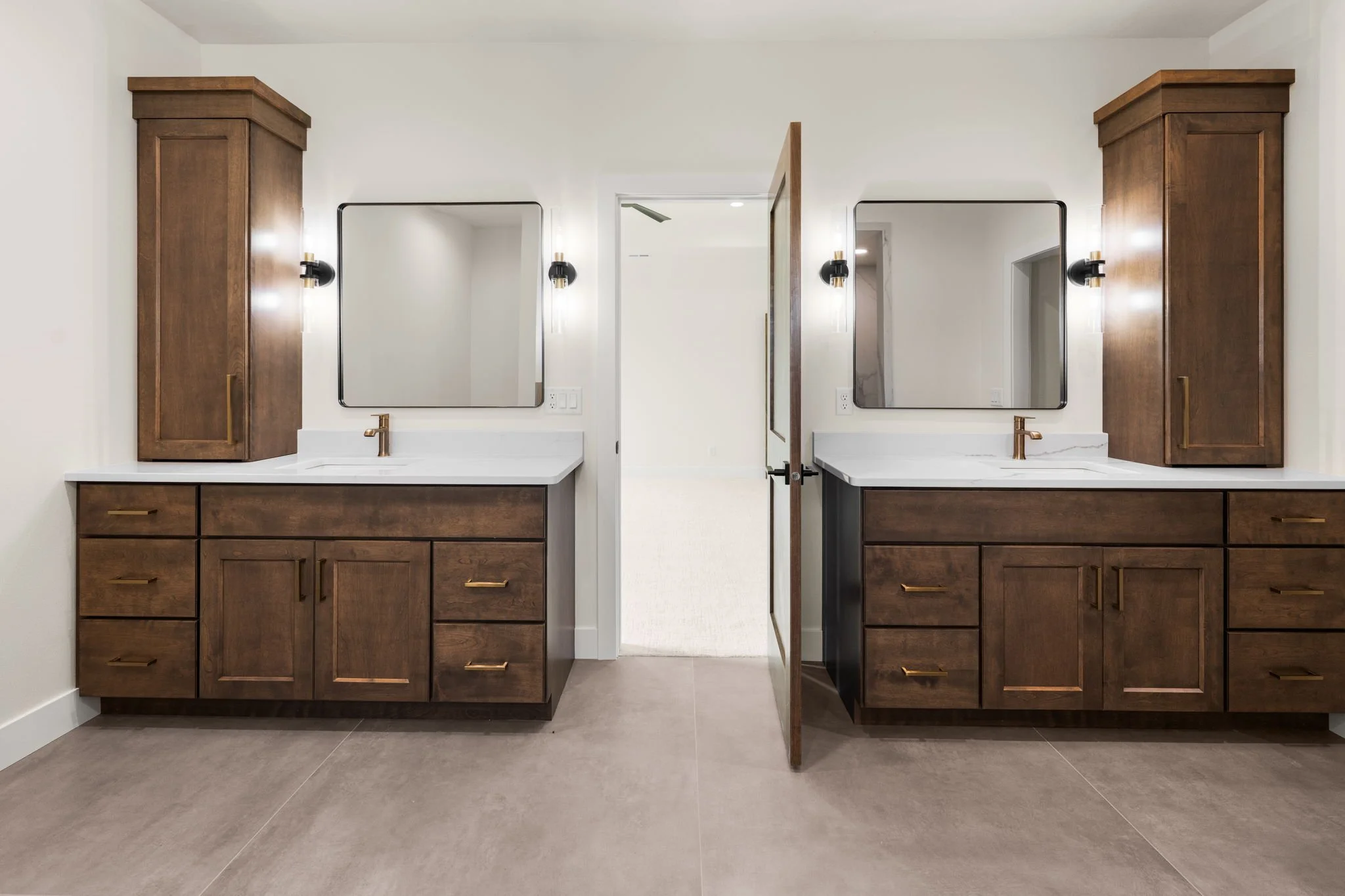 Double vanity bathroom with wooden cabinets, white countertops, and large rectangular mirrors. There are wall-mounted black and brass light fixtures above each mirror. The image shows a doorway leading to another room, with a ceiling fan visible through the doorway.