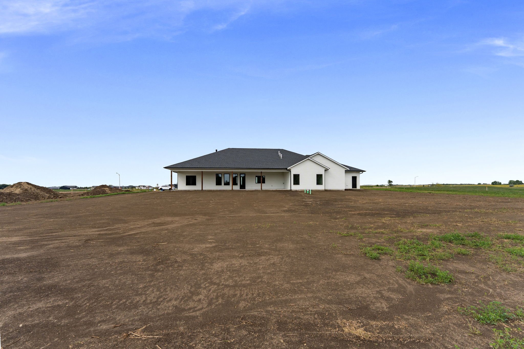 A house under construction with a grey roof and white exterior, situated on a large plot of bare dirt with patches of grass, under a blue sky.