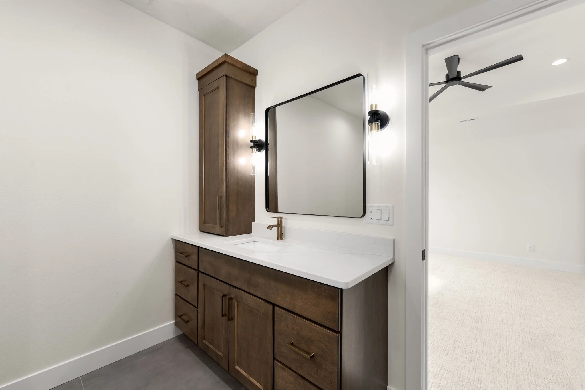 Modern bathroom vanity with a white countertop, wood cabinets, a wall-mounted mirror, black light fixtures, and a black ceiling fan in the adjacent room.