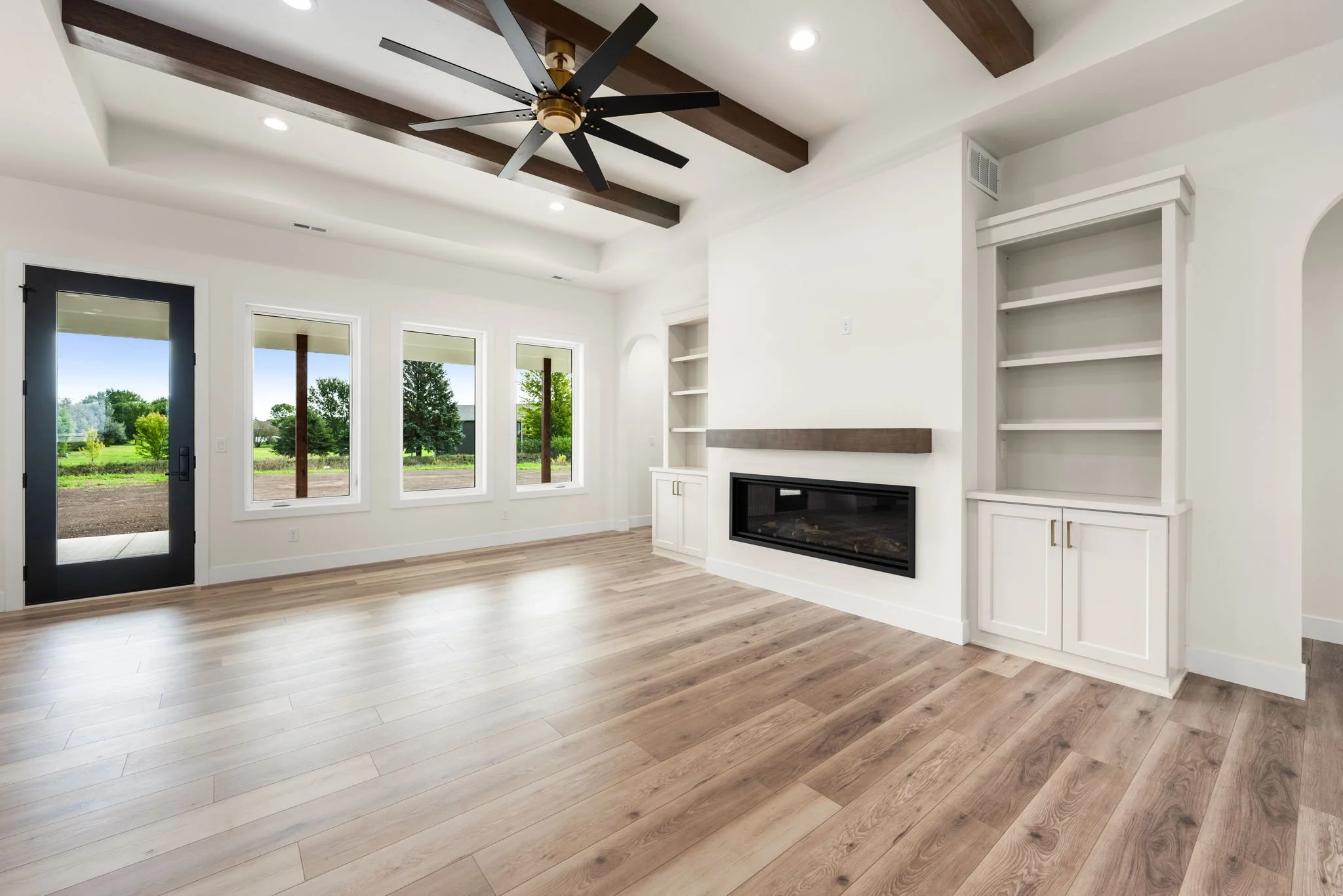 Interior view of a modern living room with white walls, large windows, a ceiling fan with black blades, a fireplace, and built-in white shelves. The room has light wood flooring and a door leading outside, with a view of greenery.