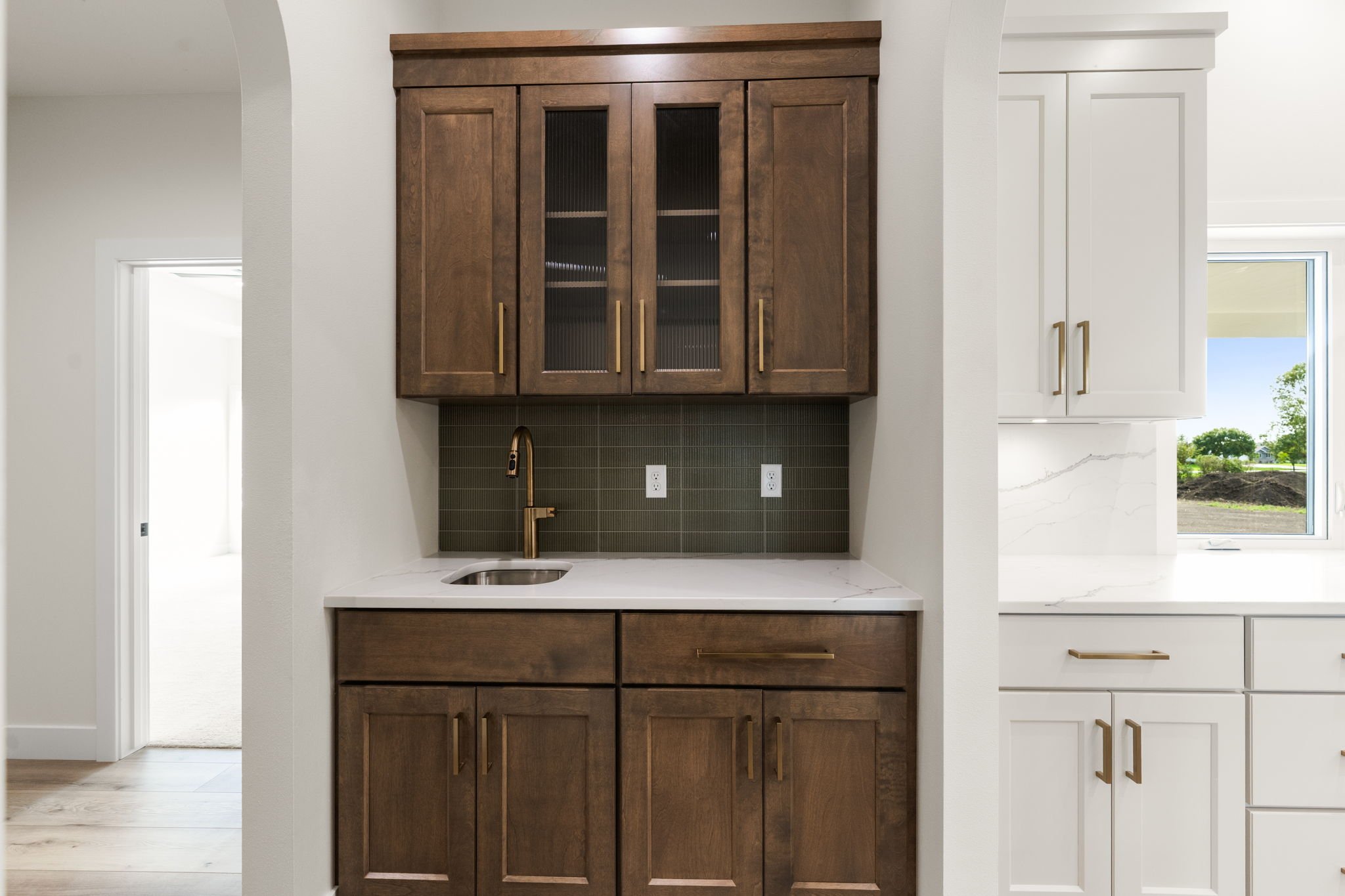 A kitchen with brown cabinets, a white countertop, a small sink, and a gold-colored faucet. There are two electrical outlets on the backsplash and a large window to the right showing trees and a blue sky.
