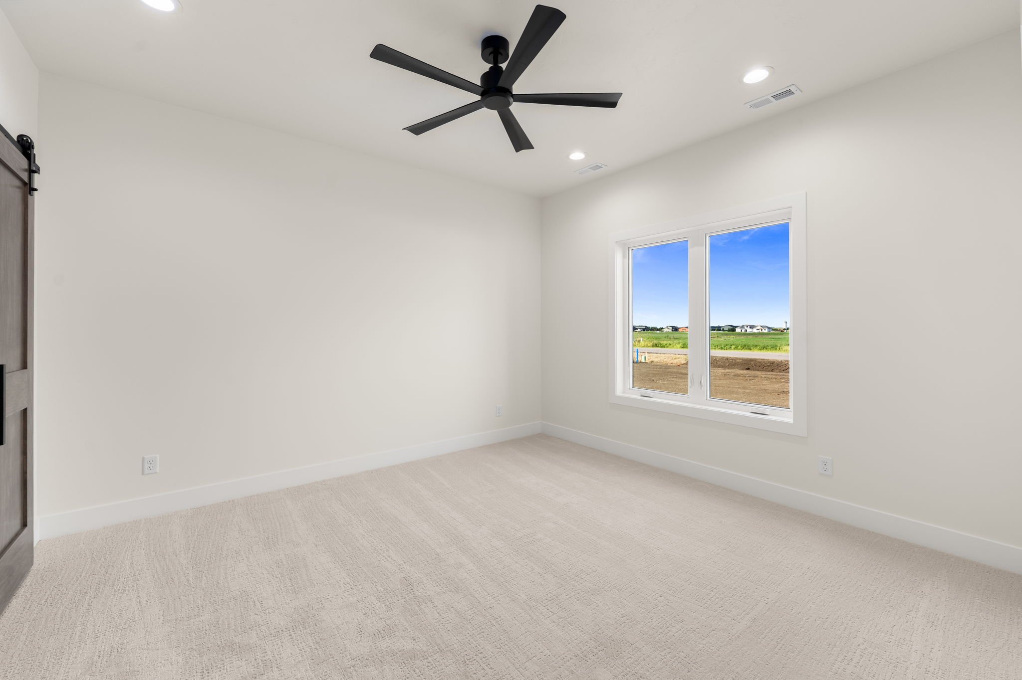 Empty room with white walls, beige carpet, a large window showing an outdoor rural landscape, a black ceiling fan, and recessed lighting.