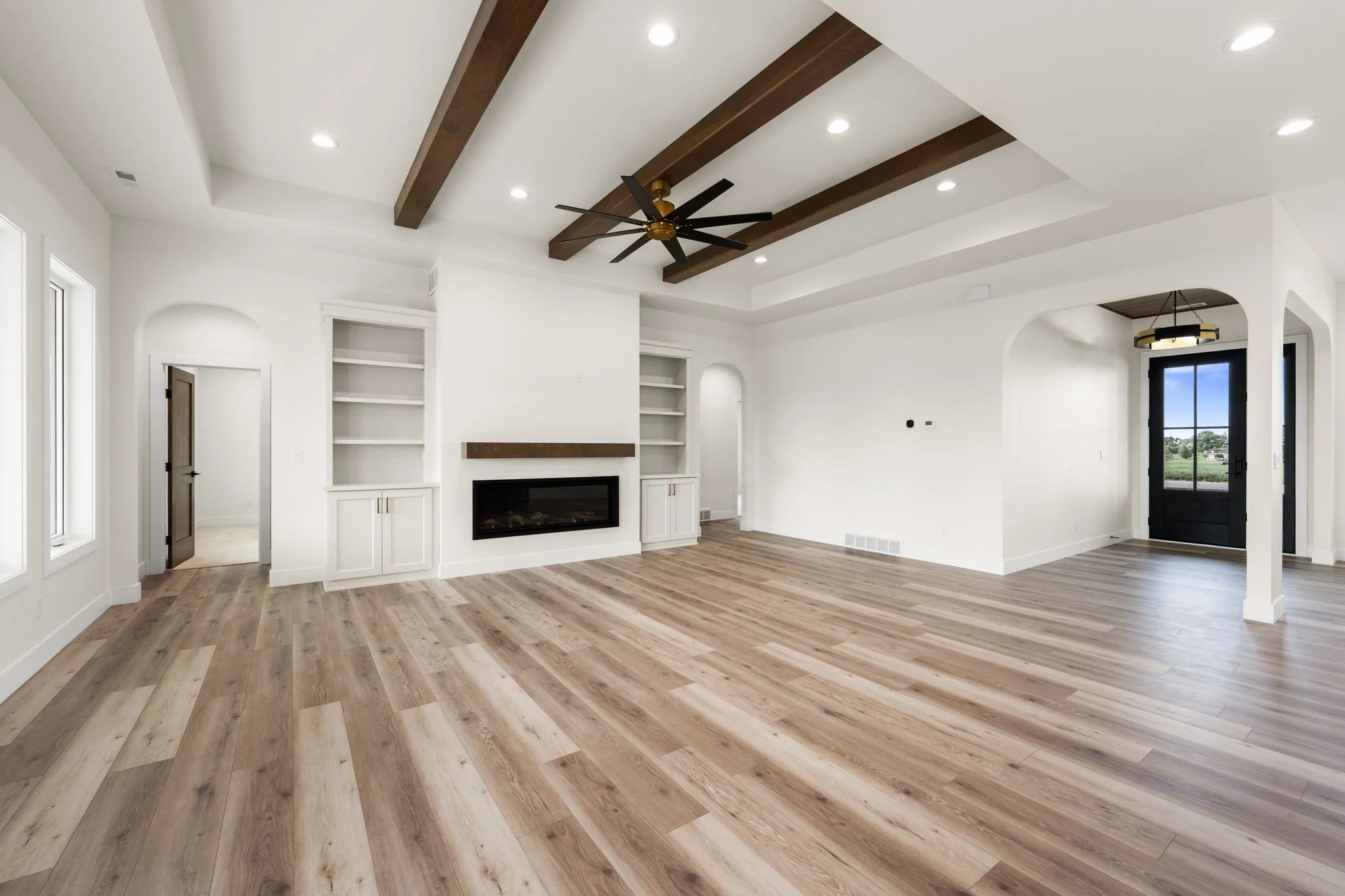 Living room with white walls, wooden beams on the ceiling, and a ceiling fan. Features built-in shelves around a fireplace, large windows, and a front door with a glass panel. Hardwood flooring.