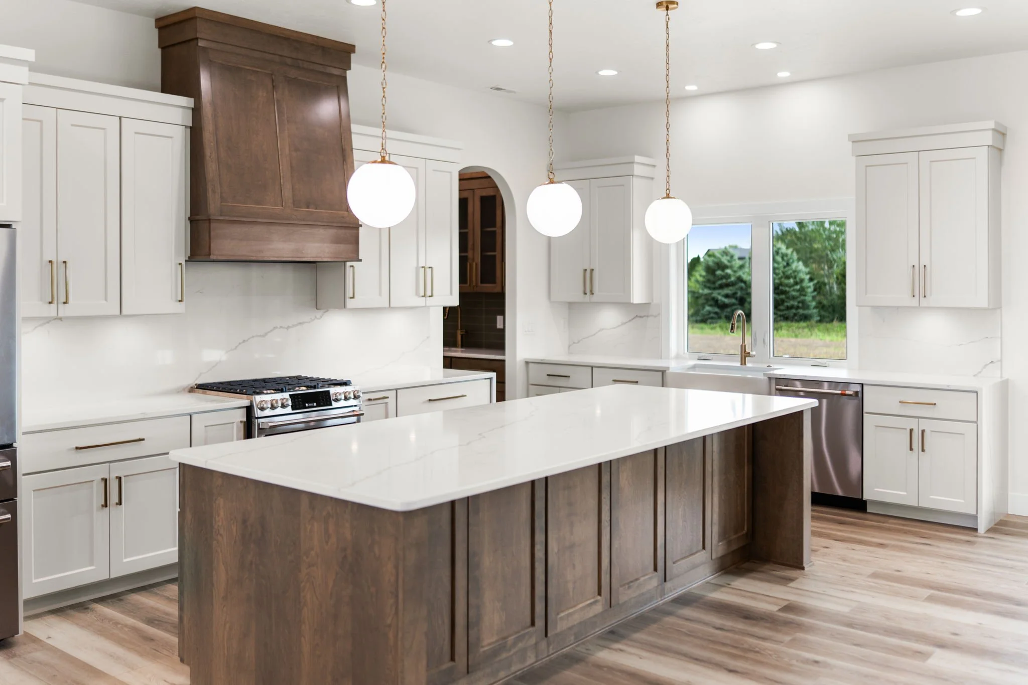 Modern kitchen with white cabinets, a large wooden island, stainless steel appliances, and three hanging globe pendant lights. Large window showing greenery outside.