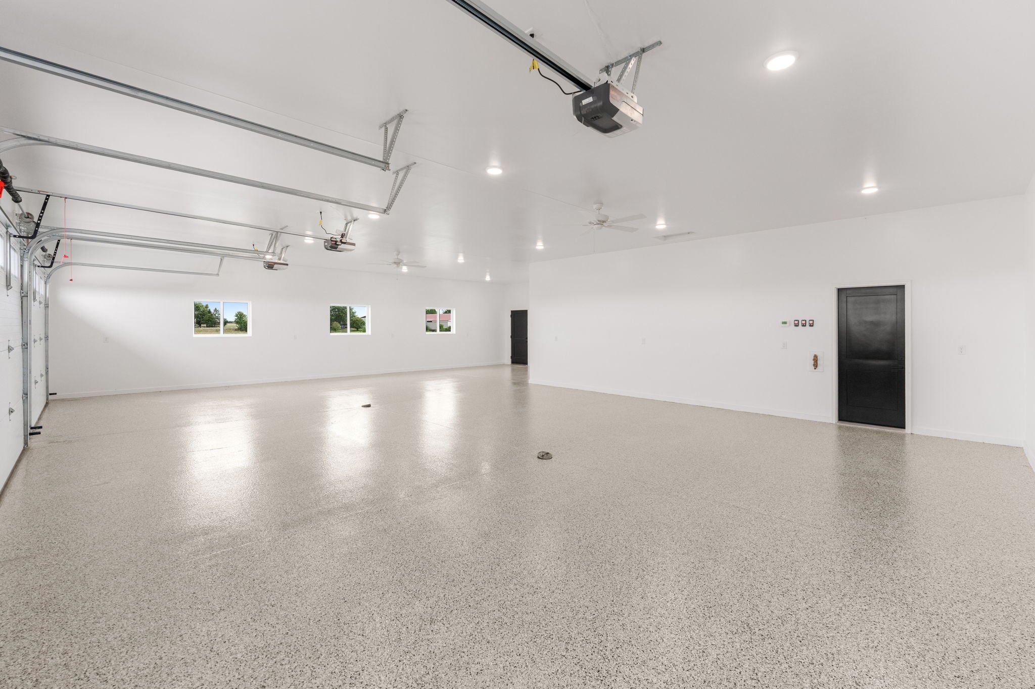 Empty garage with light-colored floor, white walls, ceiling-mounted garage door openers, and three small windows showing trees outside.