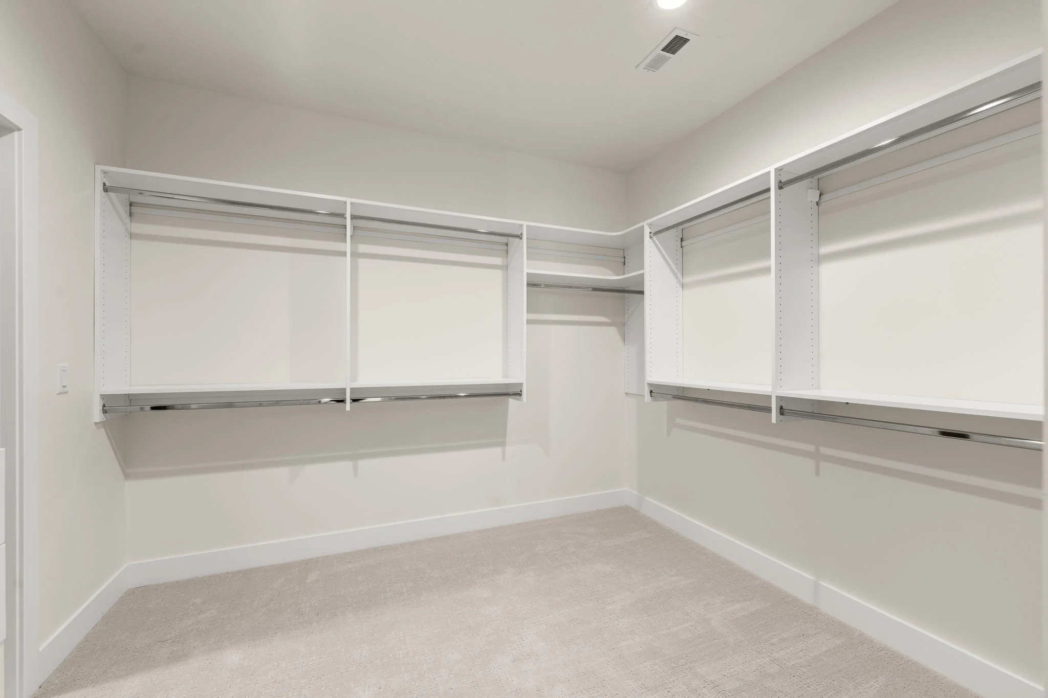 Empty white walk-in closet with shelves and hanging rods, beige carpet, white walls, and ceiling with recessed lighting and vents.