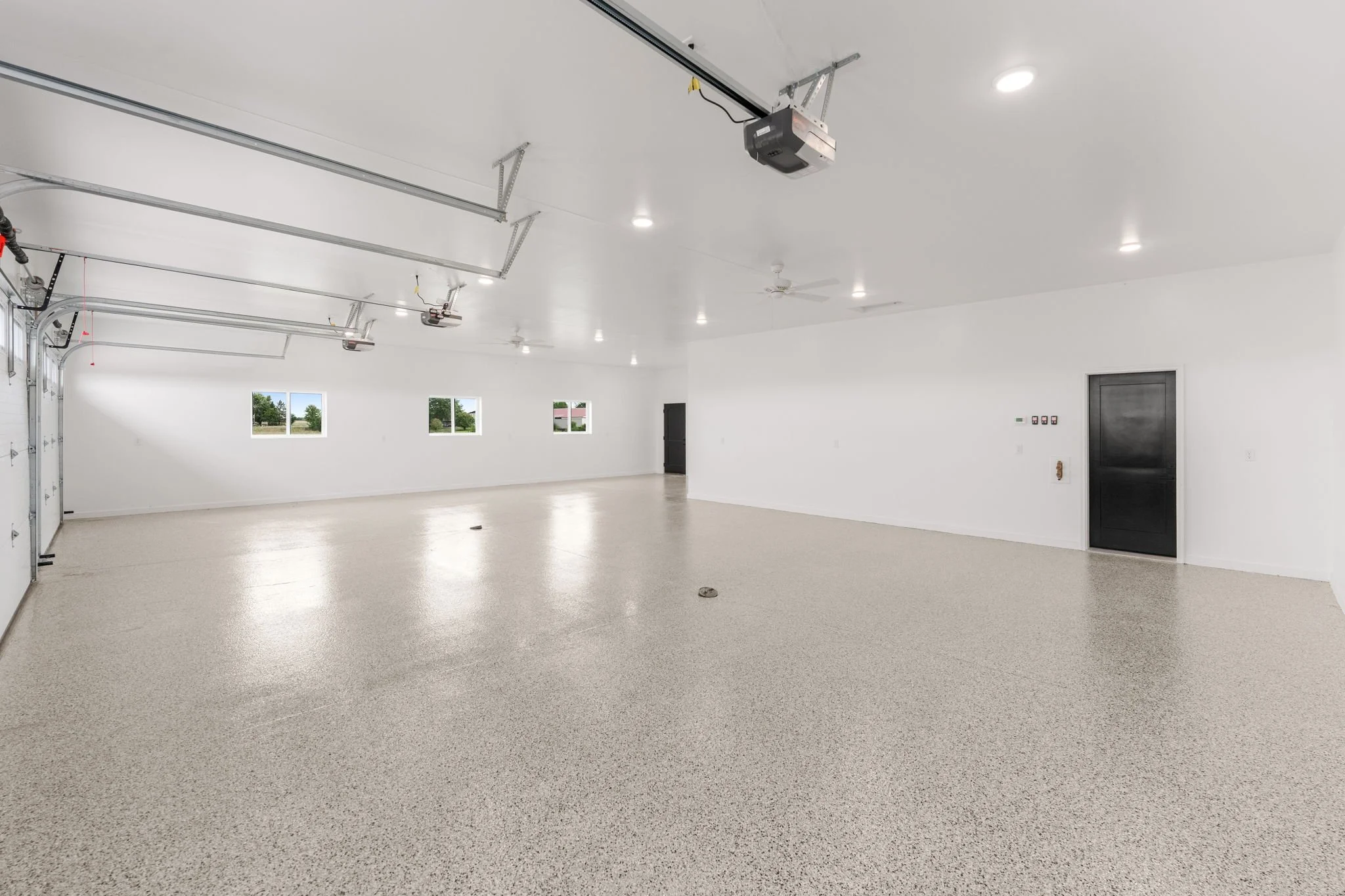 Empty garage with white walls, three small windows, a black door, and ceiling-mounted garage door openers.