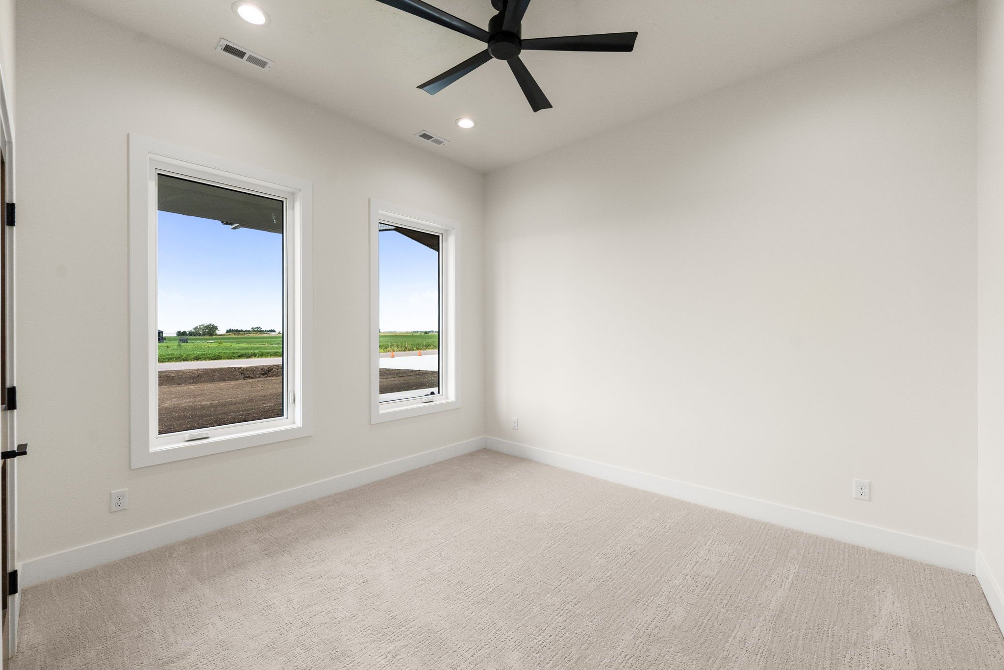 Empty room with two large windows, beige carpet, white walls, black ceiling fan, and ceiling lights.