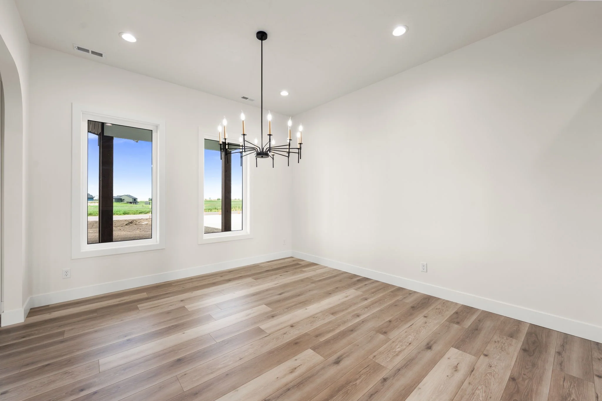 Empty room with white walls, hardwood flooring, two windows, and a black chandelier hanging from the ceiling.