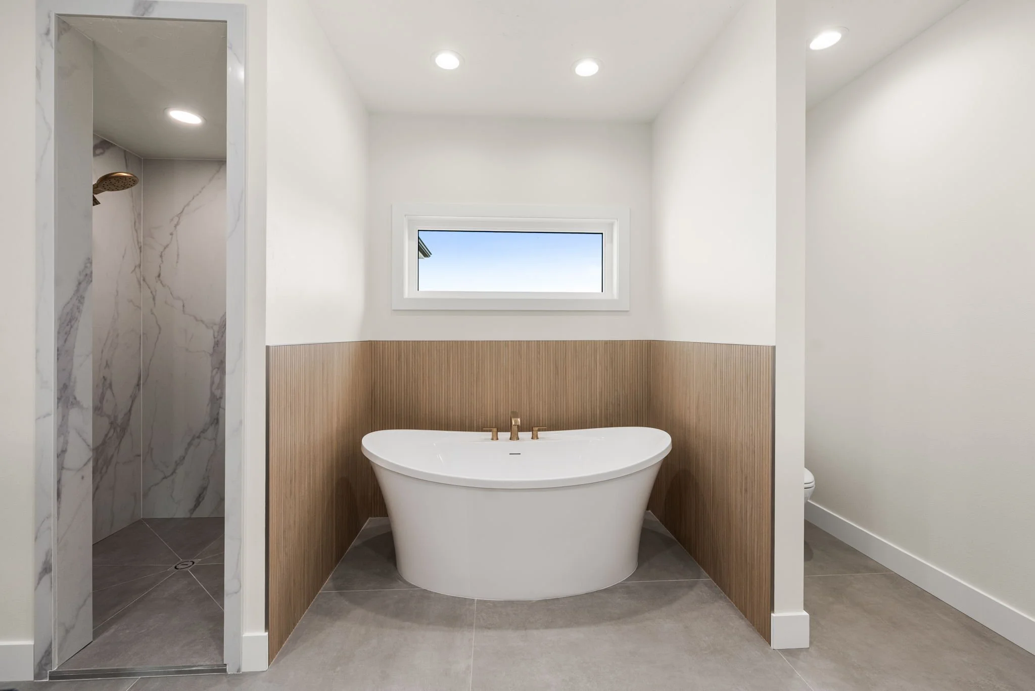 Modern bathroom with a freestanding bathtub below a window, a walk-in shower with marble walls, and neutral gray and beige tones.