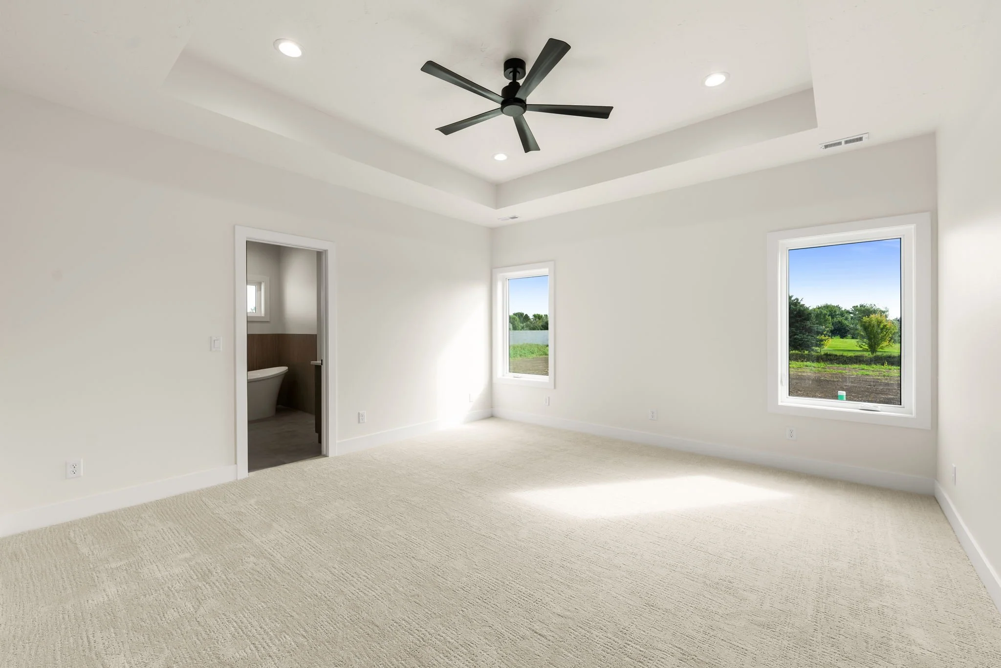 Empty room with beige carpet, white walls, three windows showing a green landscape, and a ceiling fan with four black blades.