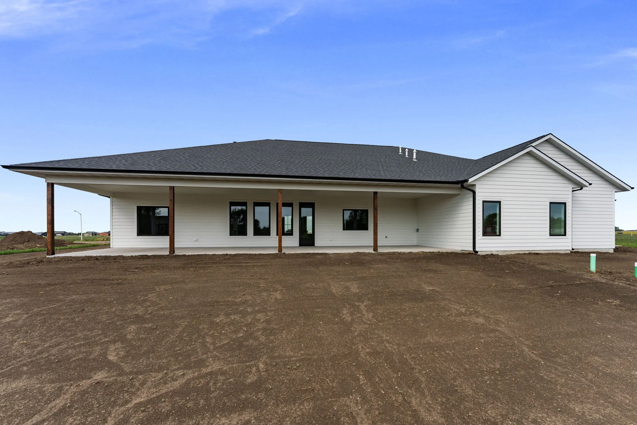 Newly constructed house with white siding, dark roof, and multiple windows, set on a dirt lot under a blue sky.