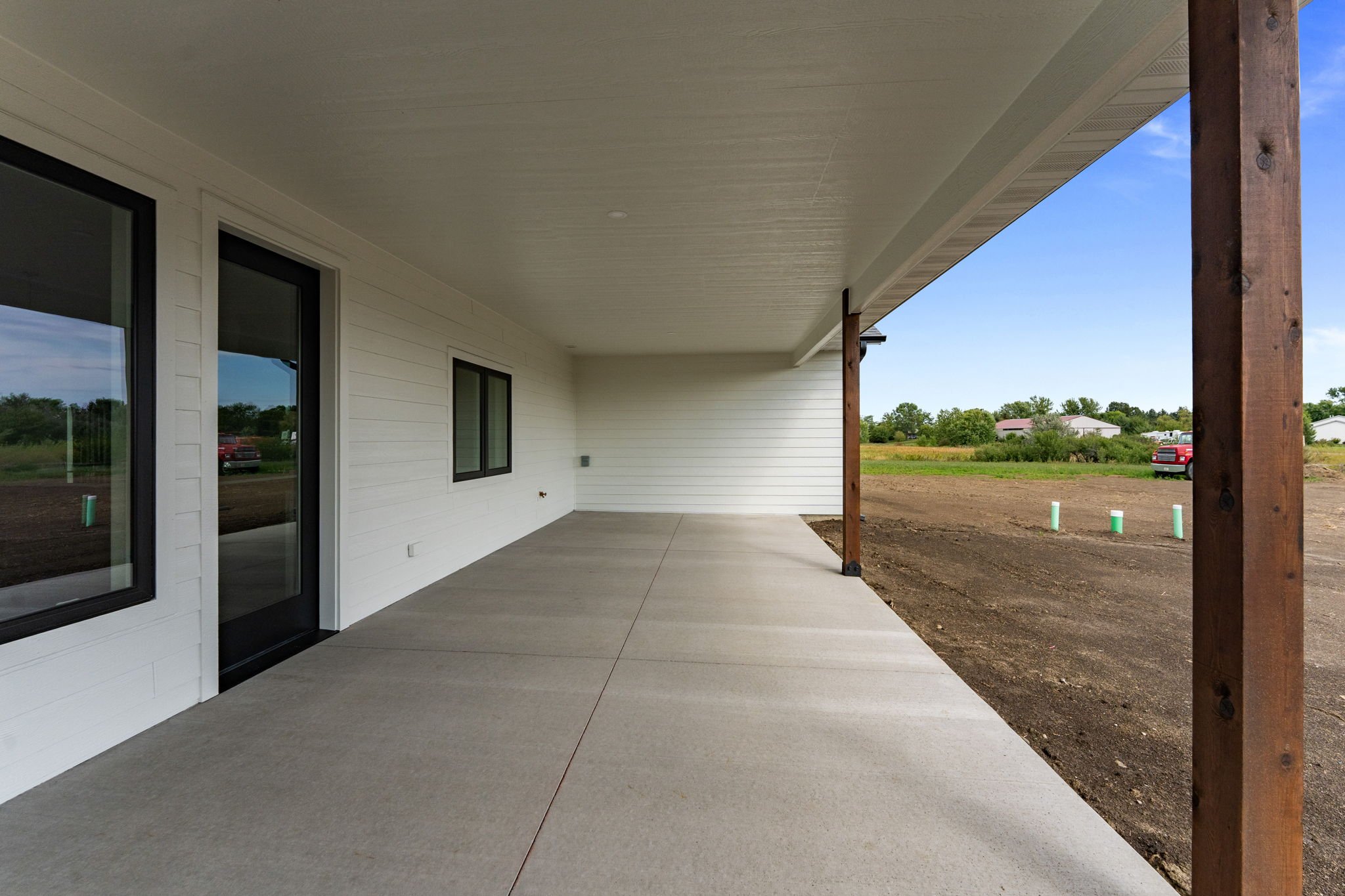 A covered porch or patio area attached to a house with white siding, black-trimmed sliding glass doors, and windows. The concrete flooring extends outward into a yard with dirt and some construction pipes. Support beams are visible, and the sky is pa