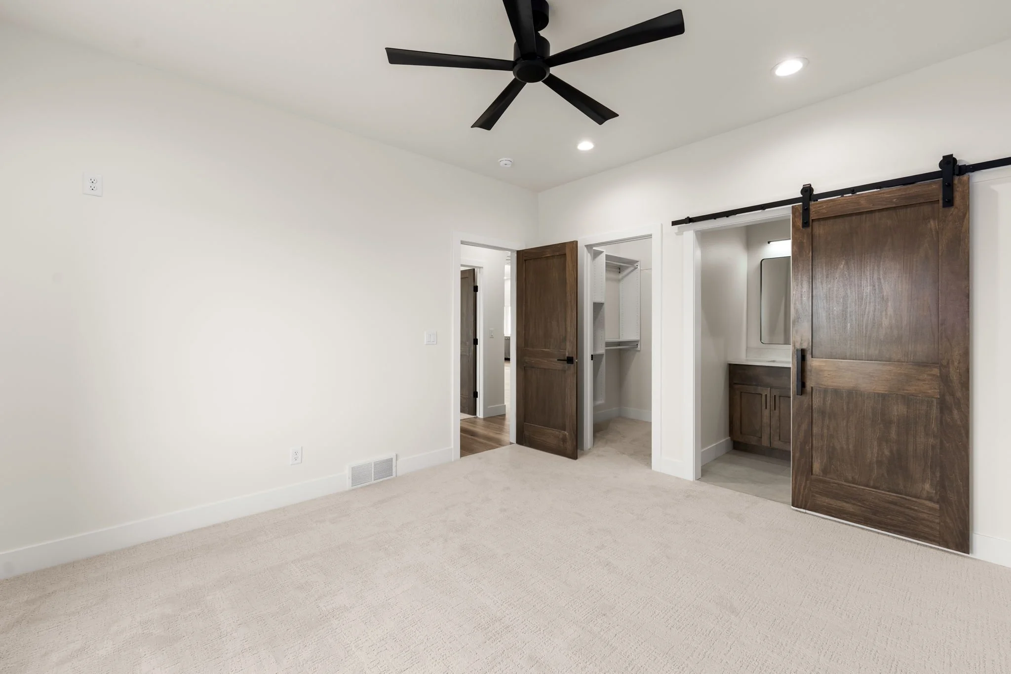 Empty room with beige carpet, white walls, four ceiling lights, a black ceiling fan, a wooden sliding barn door, an open closet with built-in shelves, and a small window in the closet.
