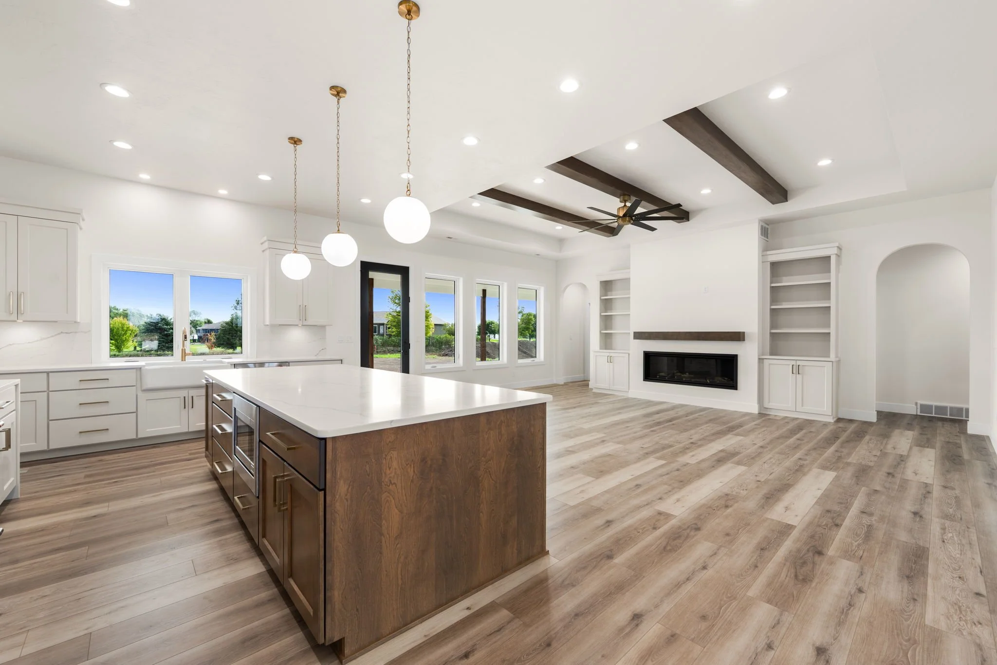 Modern open-concept kitchen and living room with white cabinetry, large island with white countertop, pendant lights, wooden floors, large windows showing greenery outside, fireplace, built-in shelves, ceiling fan, recessed lighting.