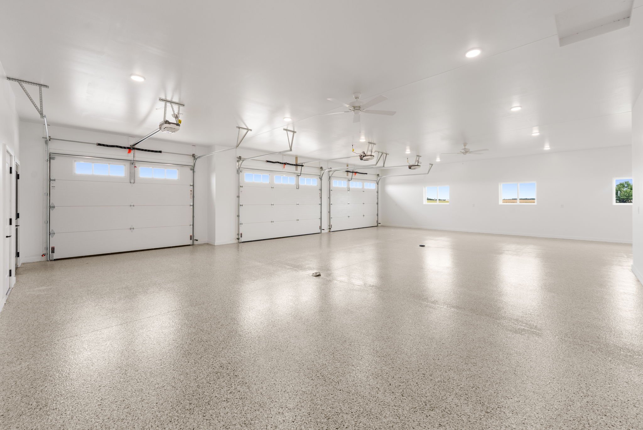 Empty spacious garage with white walls, ceiling, and garage doors, with multiple windows, ceiling fans, and ceiling lights.