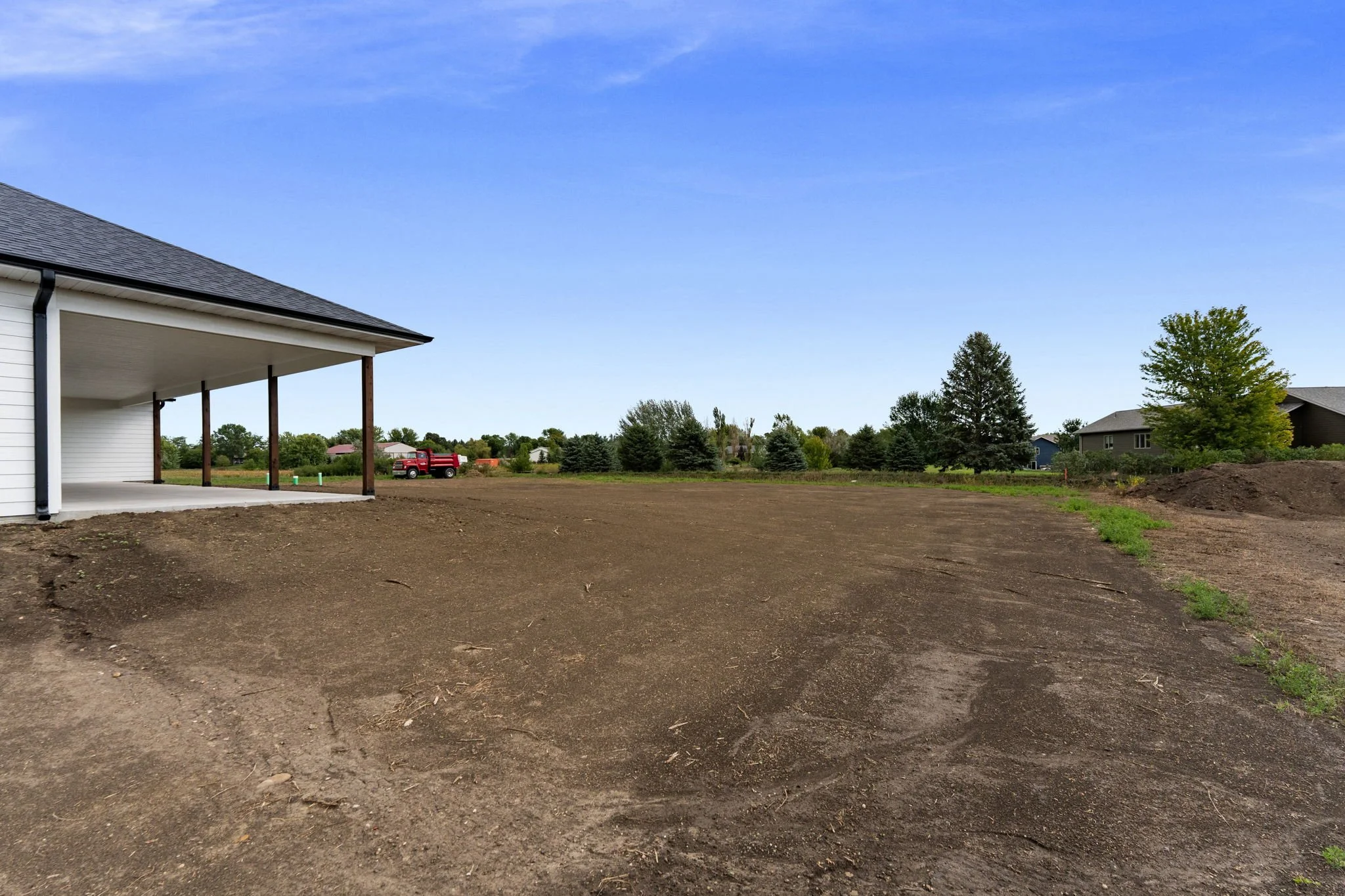 Partially constructed porch area with support beams on a house, with dirt ground and neighboring houses with trees in the background under a blue sky.