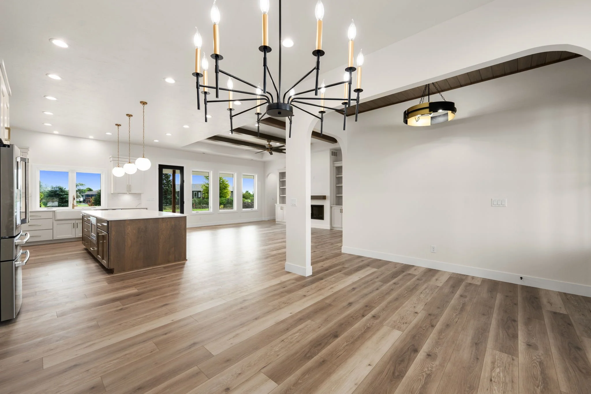 Open-concept kitchen and living area with wood flooring, white walls, large windows, and a modern black chandelier with candle-shaped bulbs hanging from the ceiling.