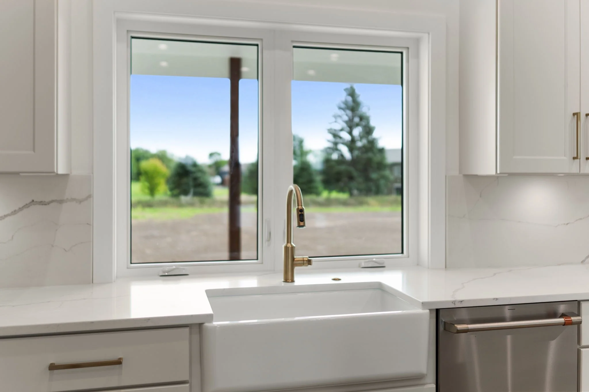 Kitchen window with a view of a green landscape, including trees and a cloudy sky, above a white marble countertop with a gold faucet and a white sink.