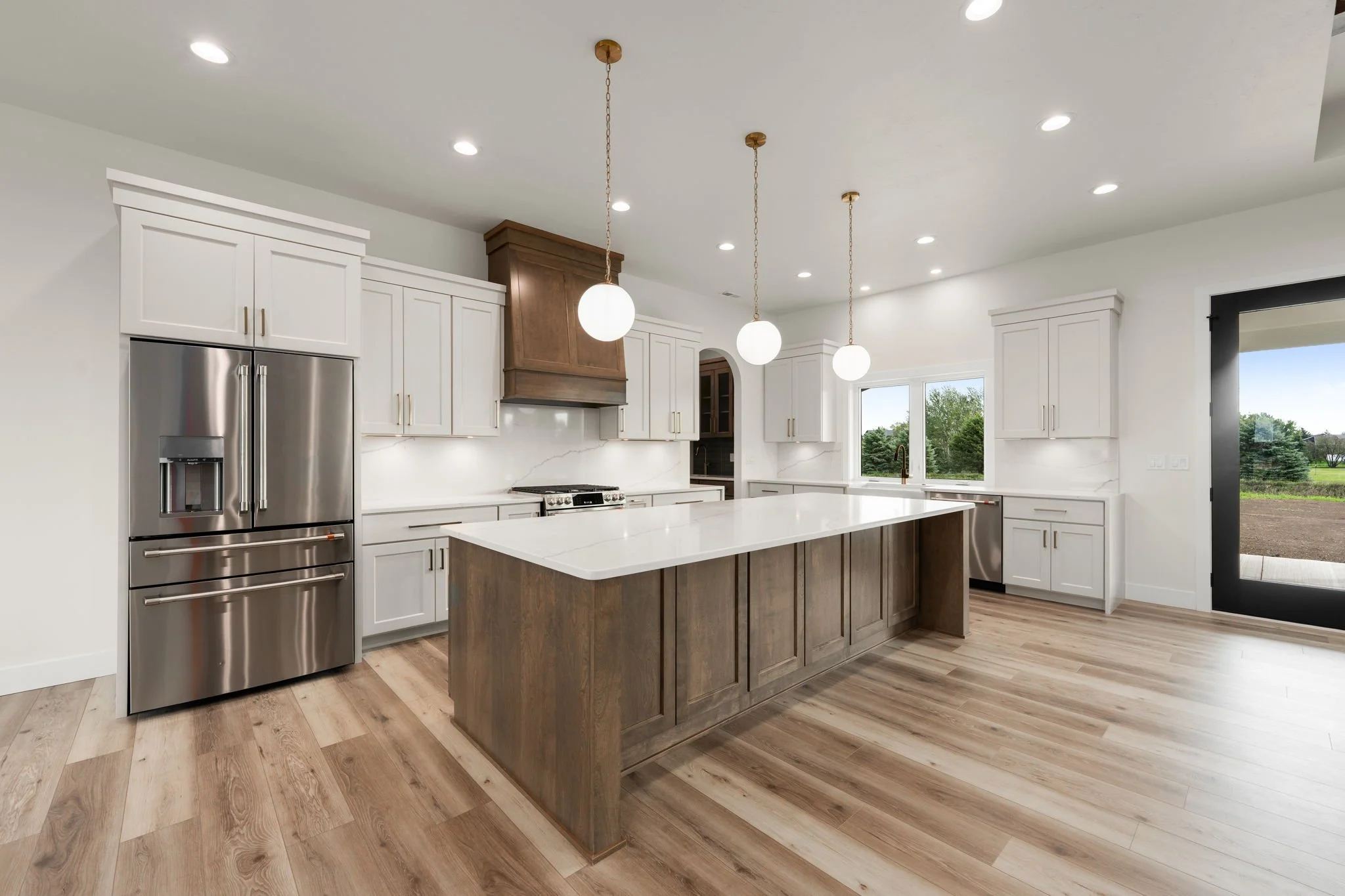 Modern kitchen with white cabinets, wooden island, stainless steel refrigerator, marble backsplash, and pendant lighting, with large windows showing outdoor greenery.