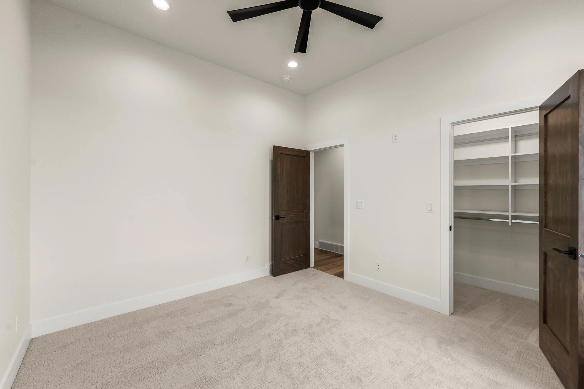Empty room with white walls, beige carpet, dark wood doors, and a ceiling fan.