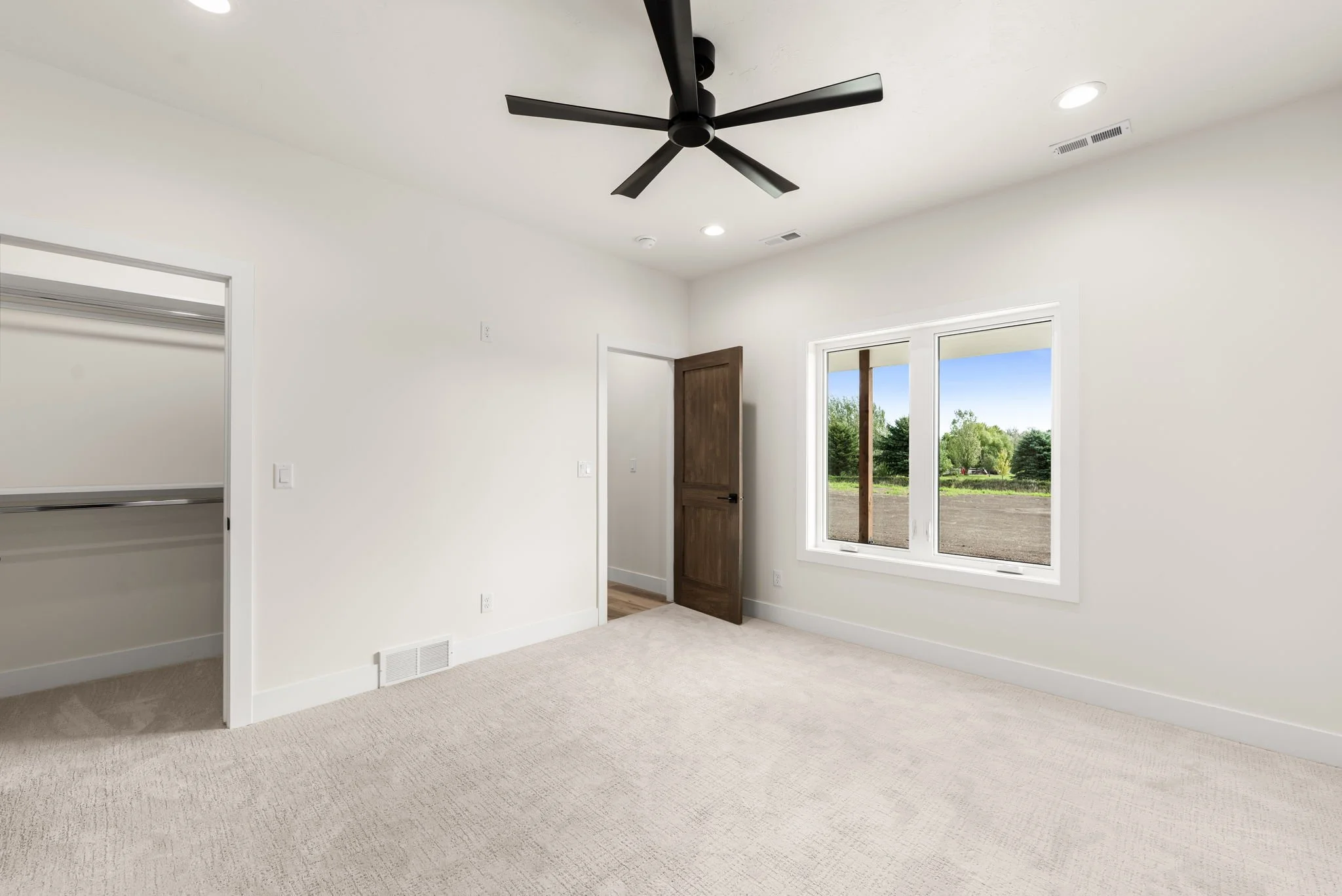 Empty room with large window showing trees and blue sky, white walls, ceiling fan, open closet, wooden door, and beige carpet.