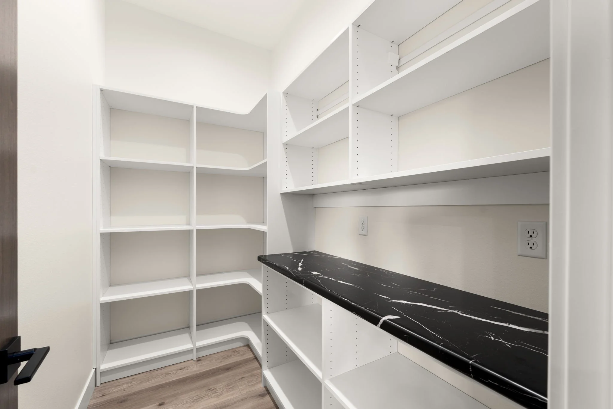 Empty white built-in corner shelves with a black marble countertop in a modern closet or pantry with hardwood flooring.