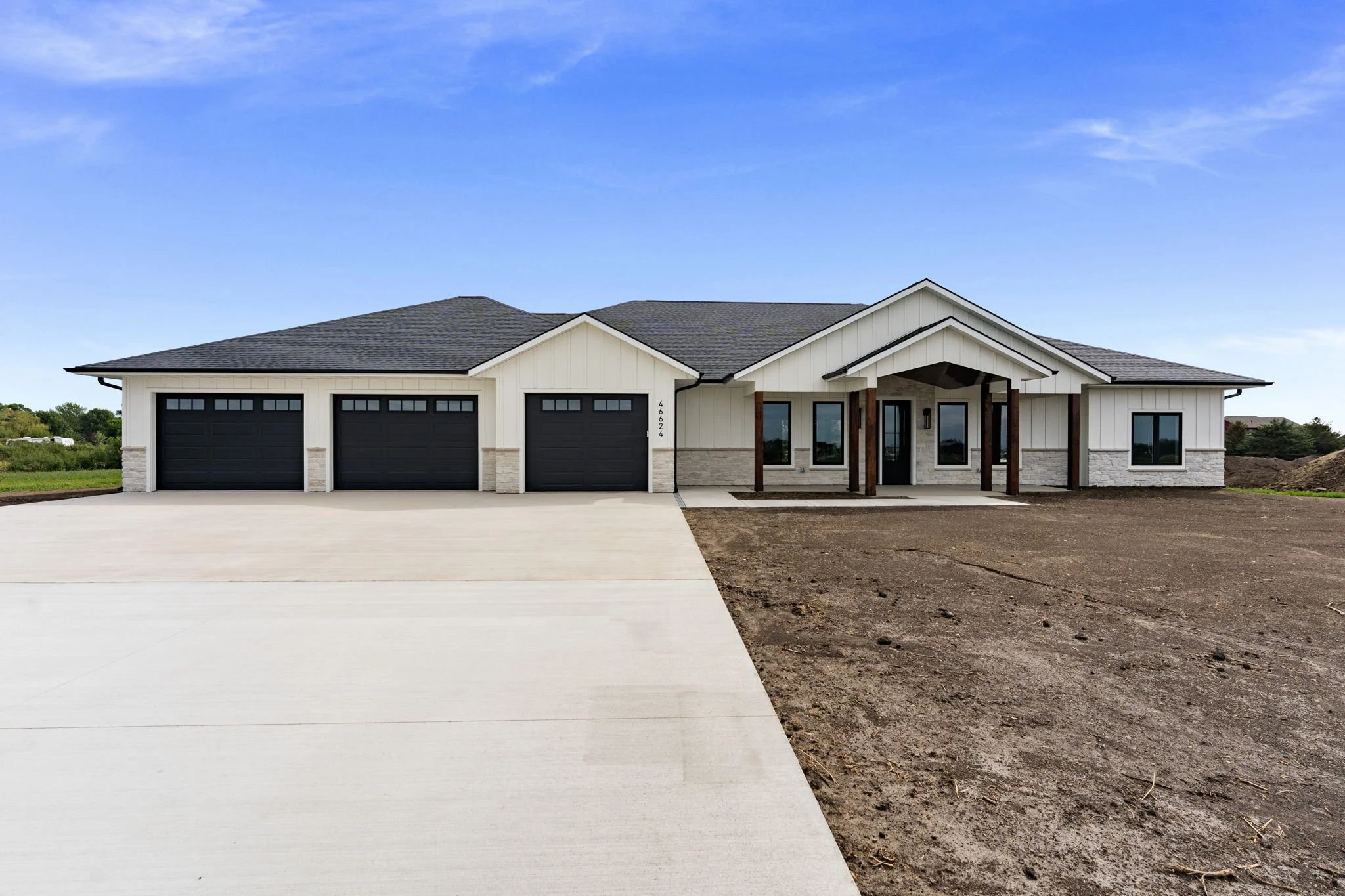 Front view of a new modern house with a gray roof, white exterior walls, and a front porch supported by wooden beams, with a driveway and an empty lot of dirt in front.