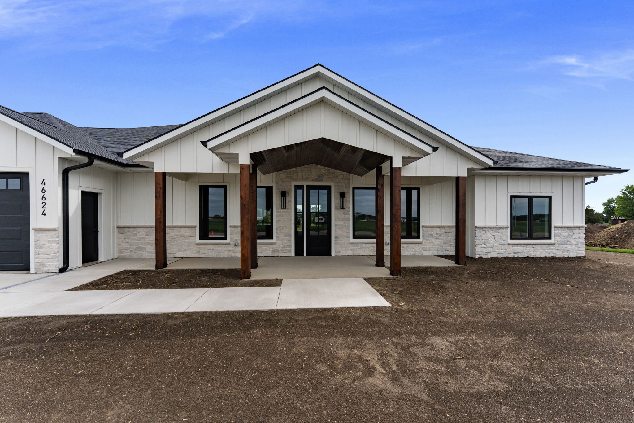 Newly constructed house with white siding, stone accents, and a covered porch supported by wooden posts, situated in a suburban area with an empty dirt yard and clear blue sky.