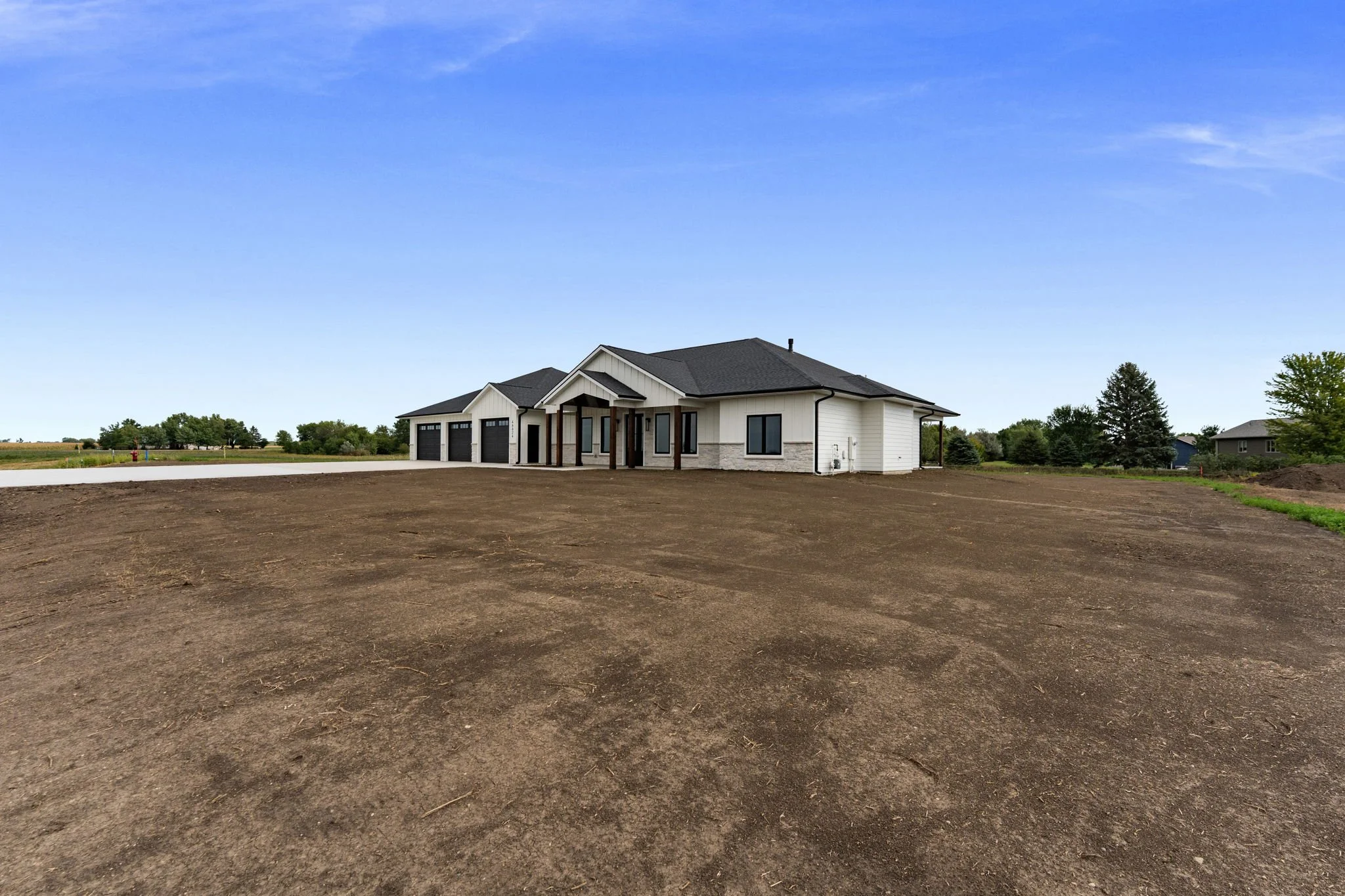New house with a large front yard under construction, featuring a two-car garage, in a rural area with trees and a blue sky.