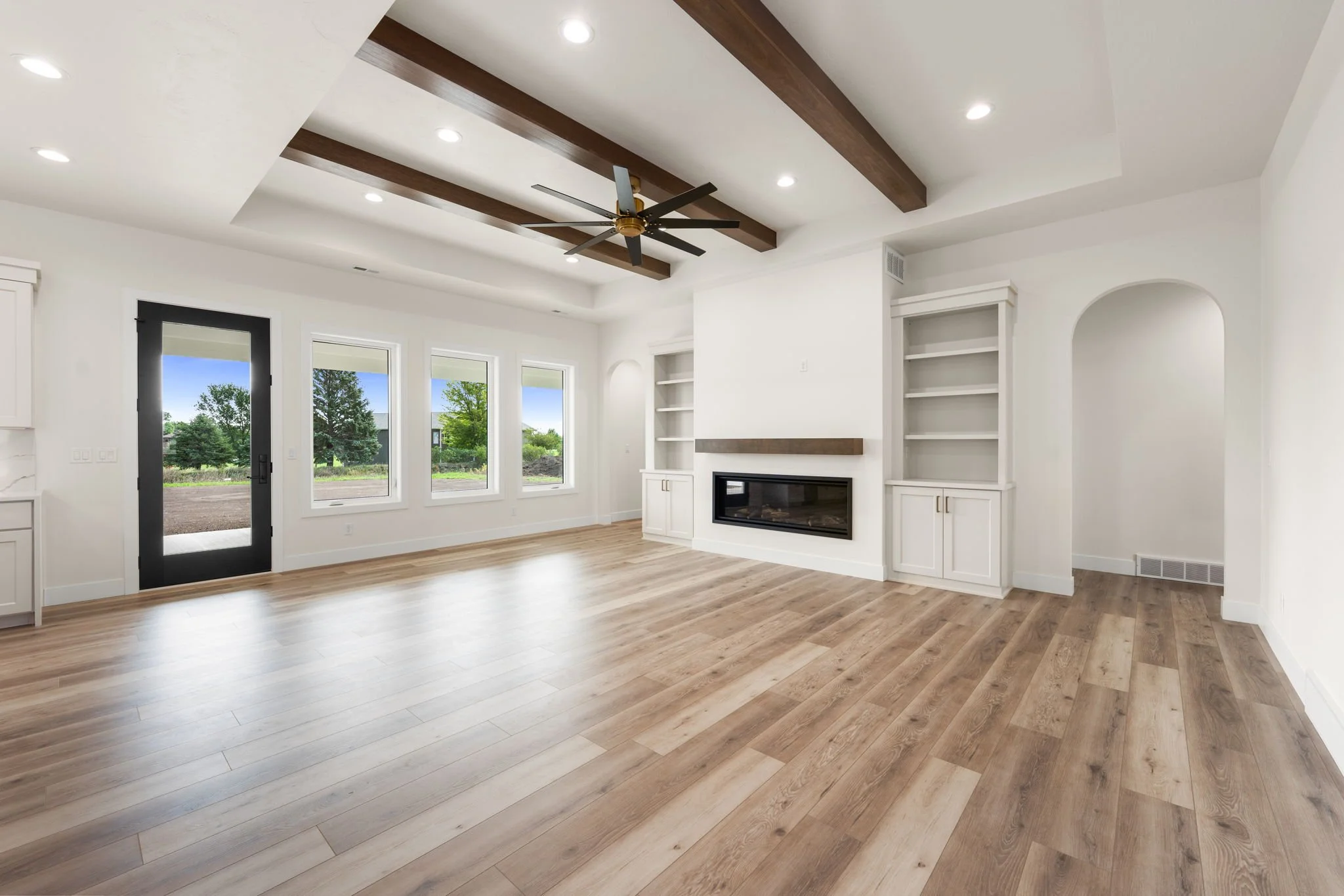 Living room with hardwood flooring, white walls, built-in shelving, a fireplace, large windows with trees outside, and a ceiling fan with wooden beams.