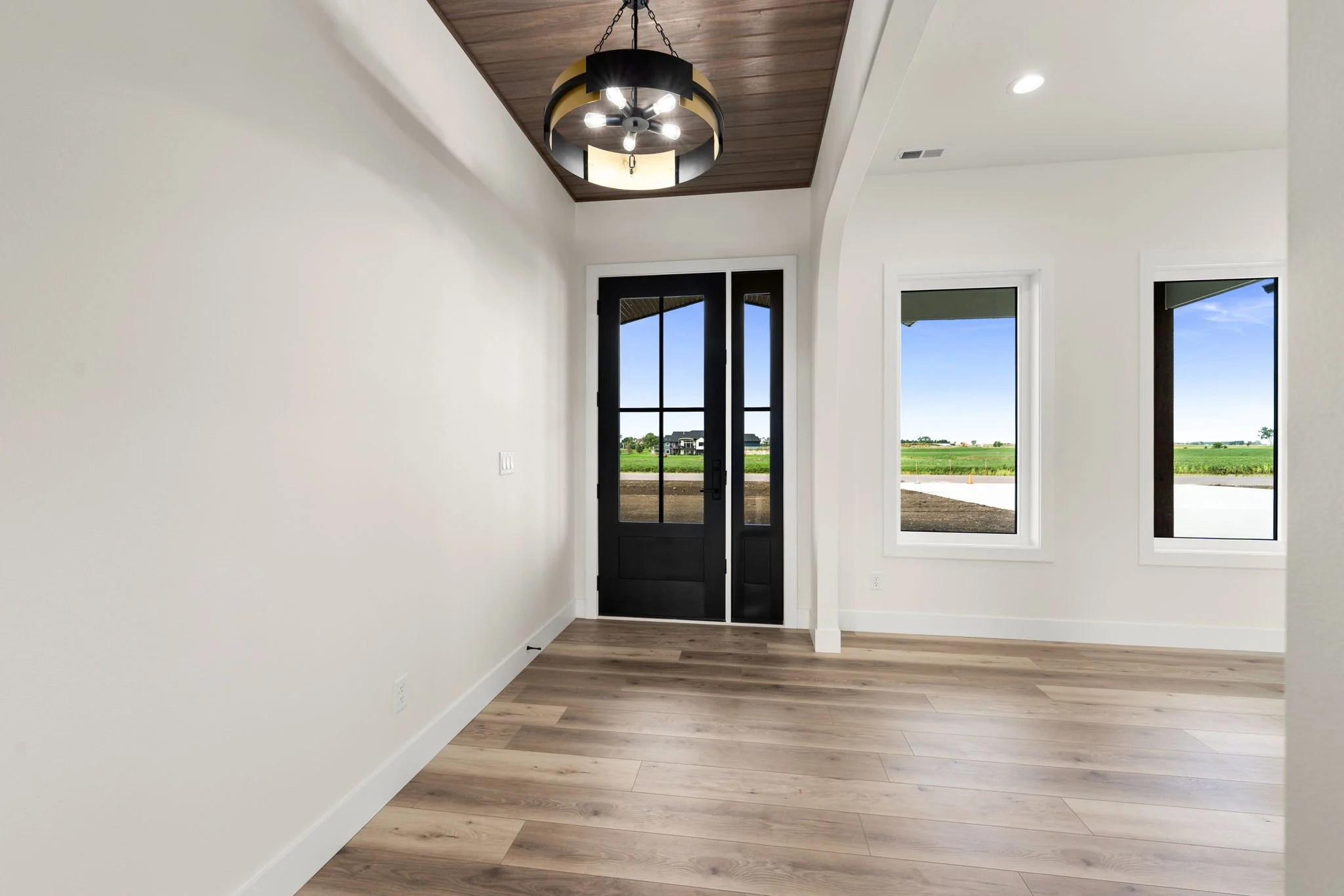 Interior view of a bright, empty room with white walls, a wooden floor, large windows, and a modern black door. The ceiling has a wooden panel and a black and gold chandelier.