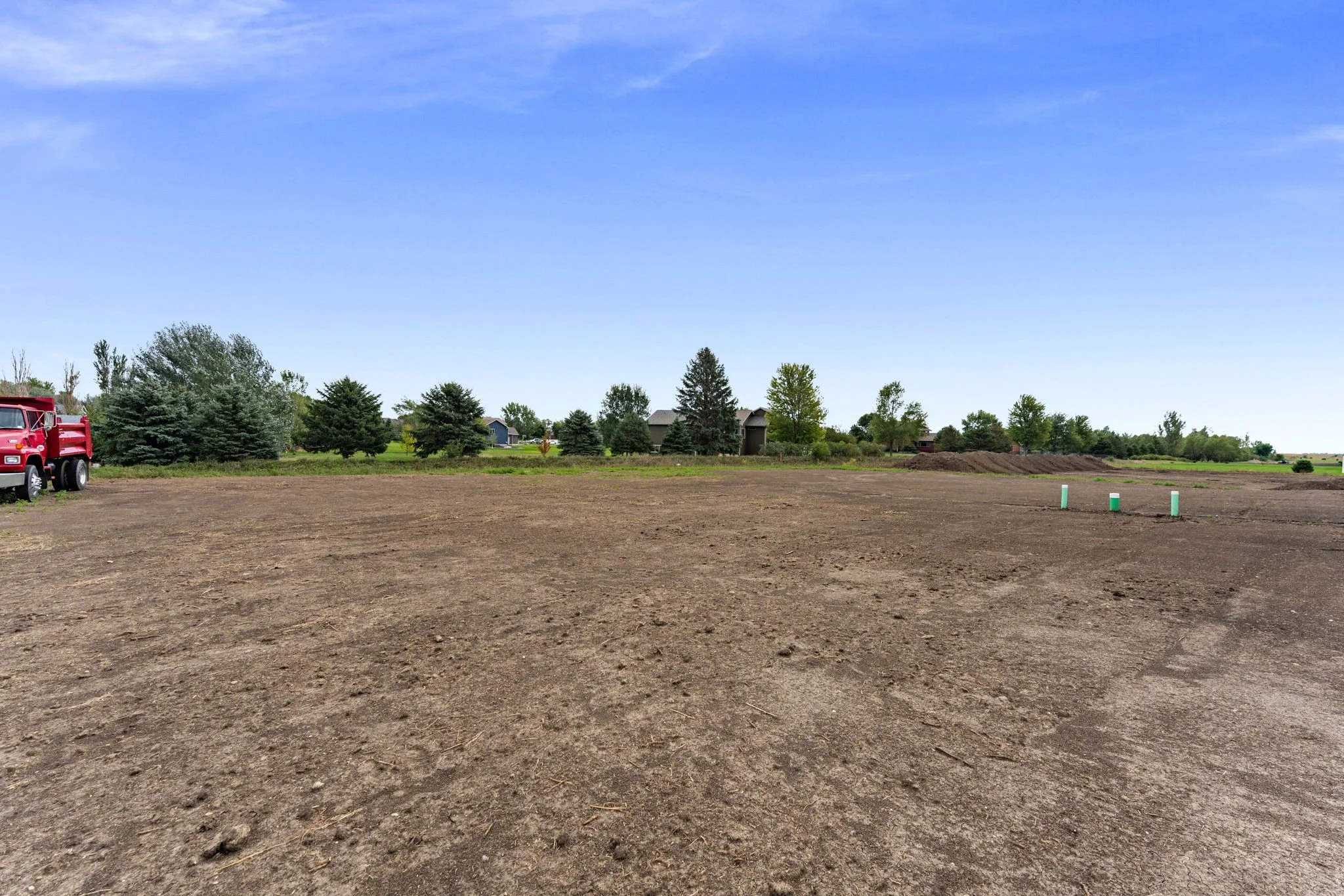 A large open dirt lot with a red truck parked on the left side and a row of trees and houses in the background under a clear blue sky.