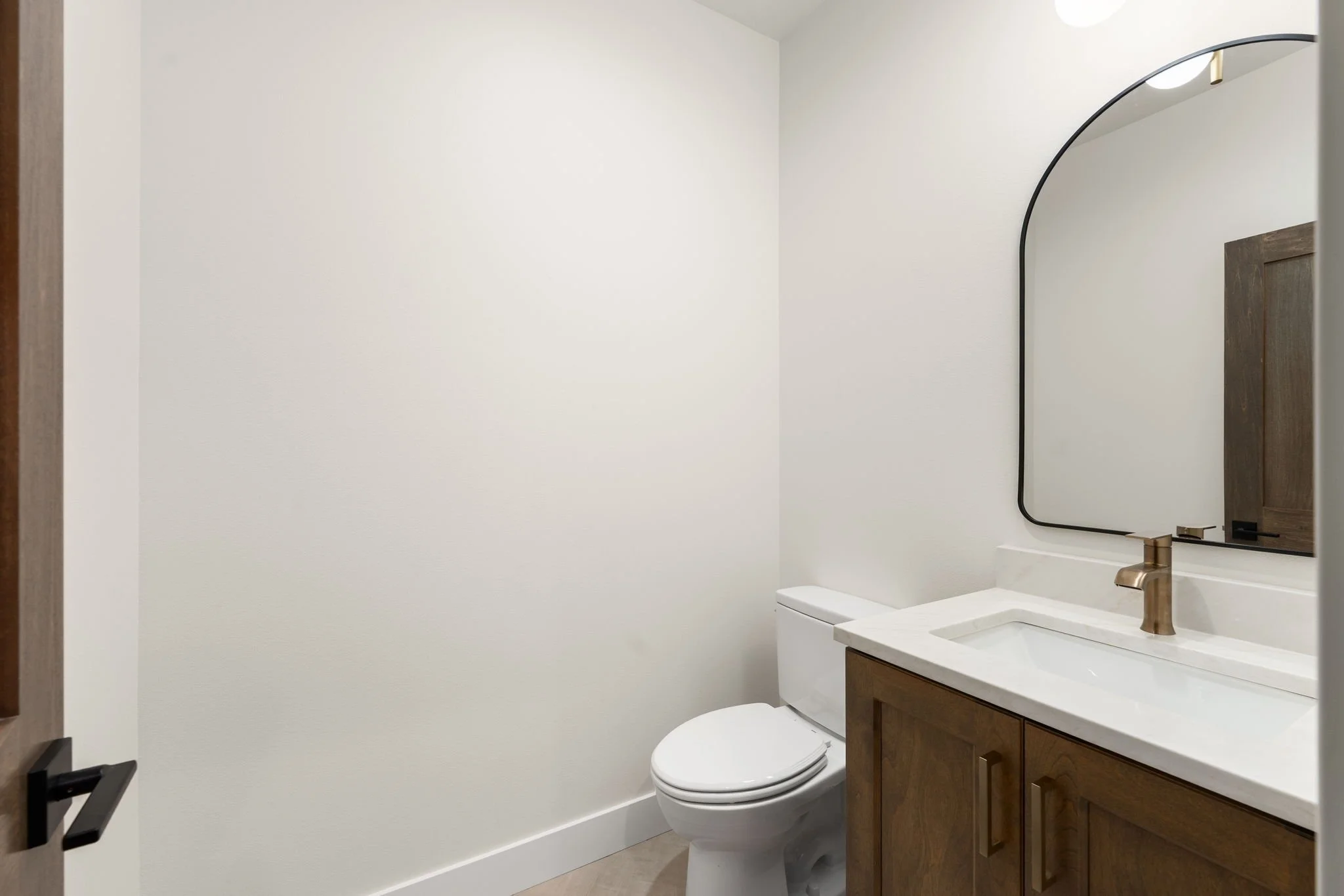 A modern bathroom with a white toilet, a white vanity with a sink, a gold faucet, a large mirror with a black frame, and wooden cabinet doors. The walls are painted white, and there is a ceiling light fixture.