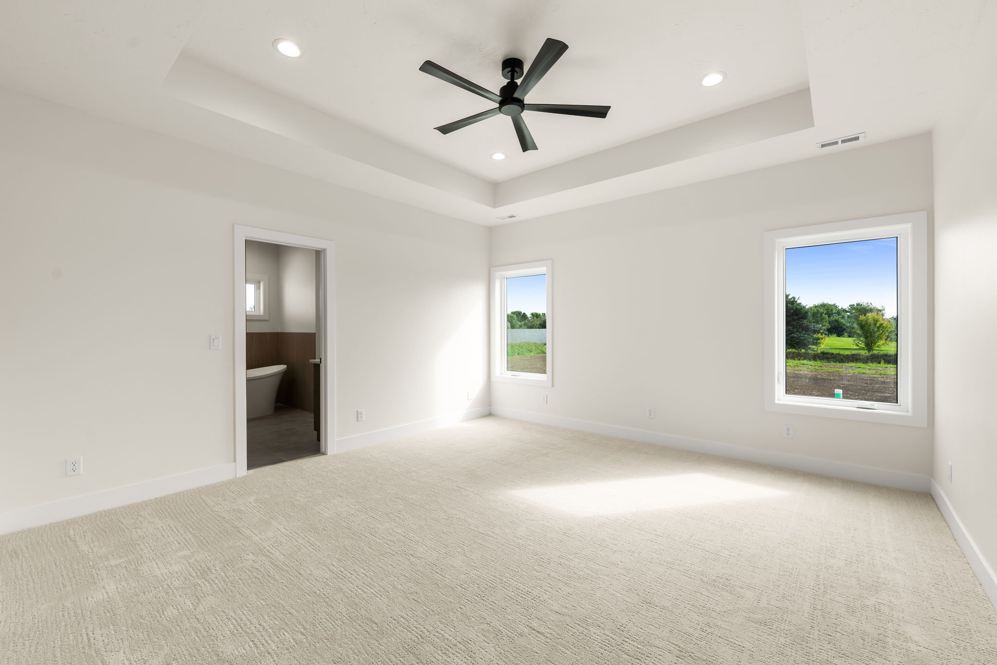 Empty living room with beige carpet, white walls, two windows showing greenery outside, ceiling fan, and recessed lighting.