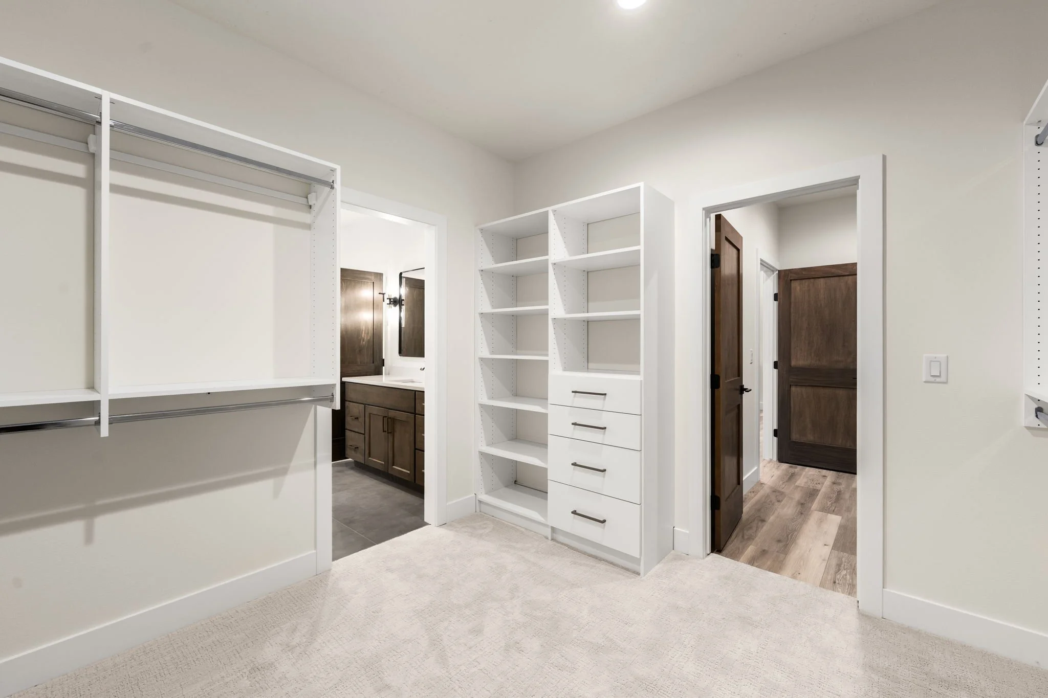 Empty walk-in closet with white shelves and hanging rods, adjacent to a bathroom with wooden cabinets and dark brown doors.