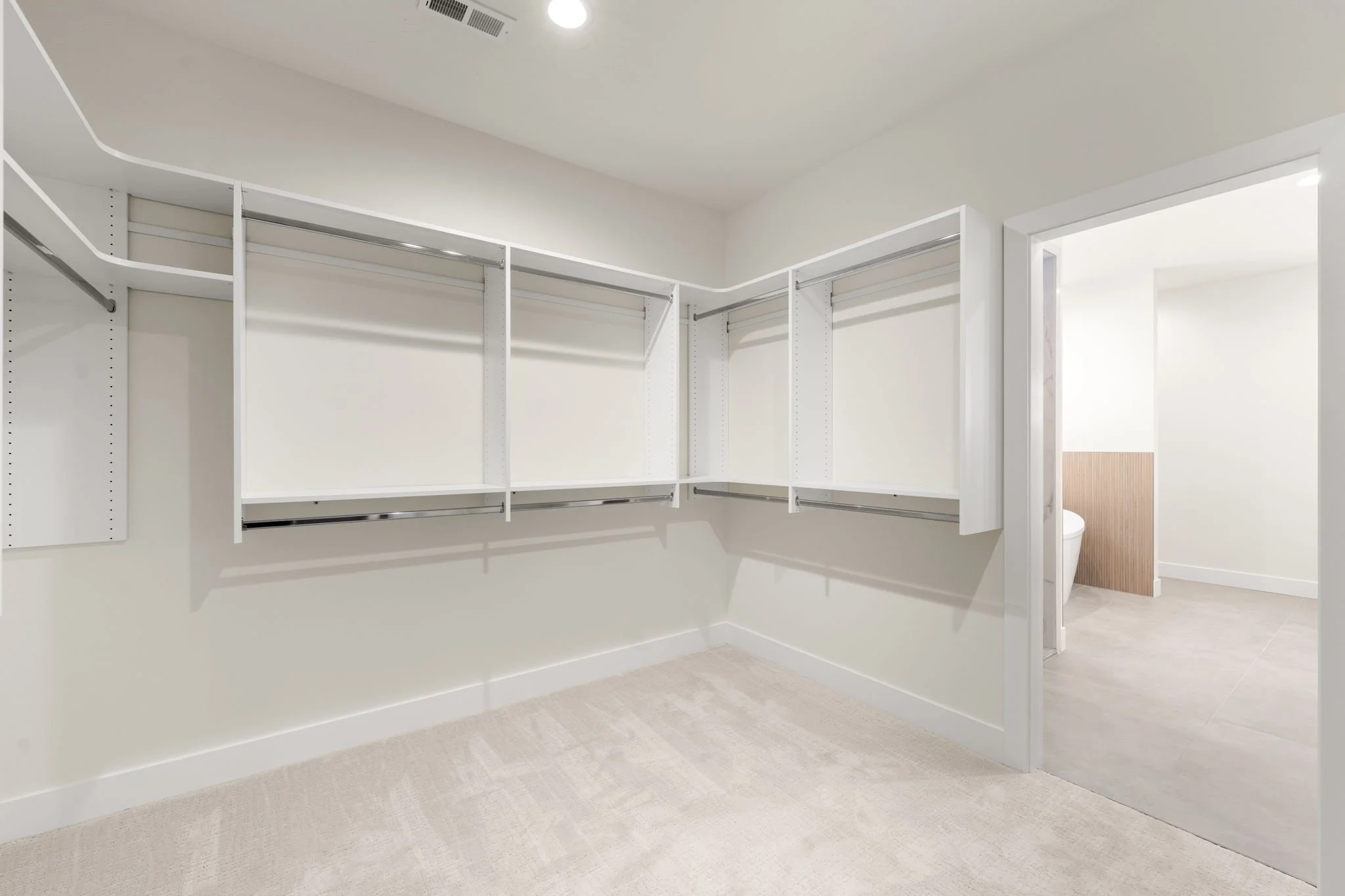 Empty walk-in closet with white shelves and rods, beige carpet, and a doorway leading to a bathroom with toilet and wood-paneled wall.